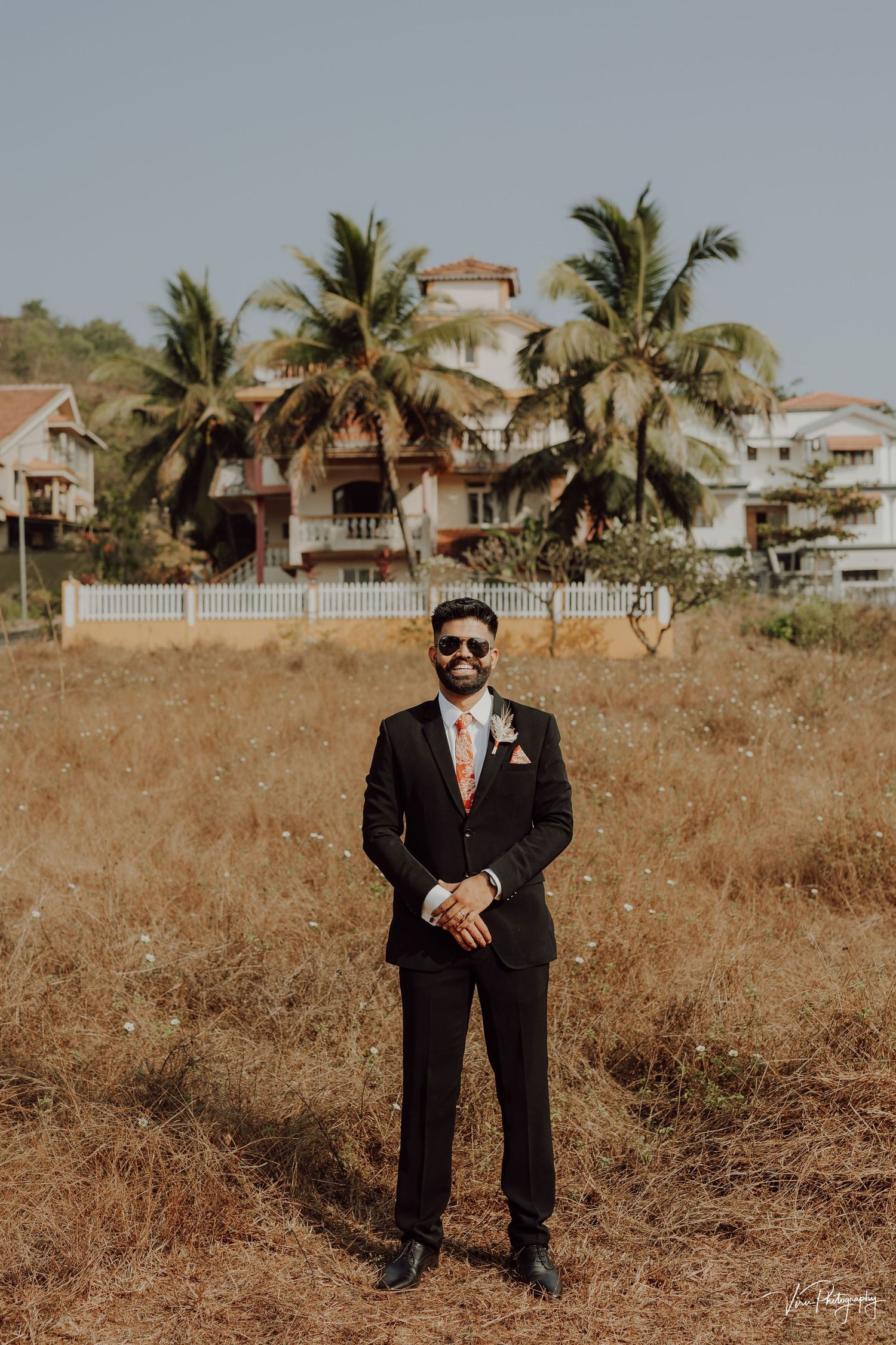 A suited figure stands in a dry field with palm trees and coastal buildings in the background on a sunny day.