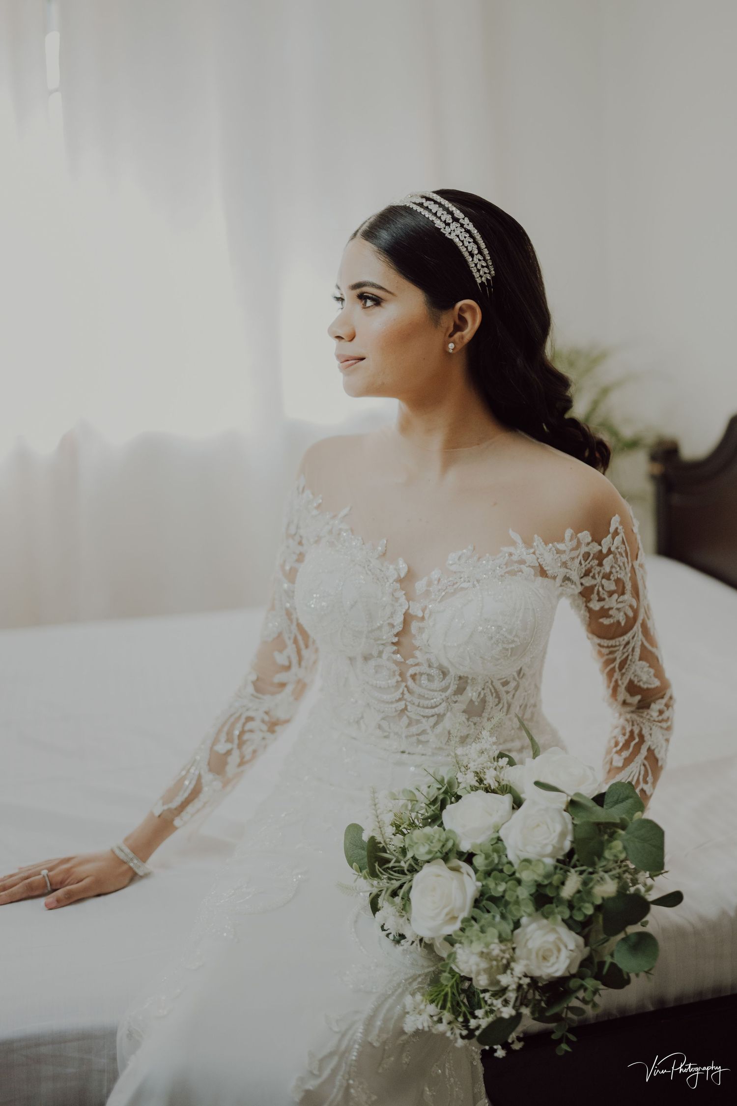 Elegant bride in long sleeve lace wedding gown holds white rose bouquet while sitting in soft window light.