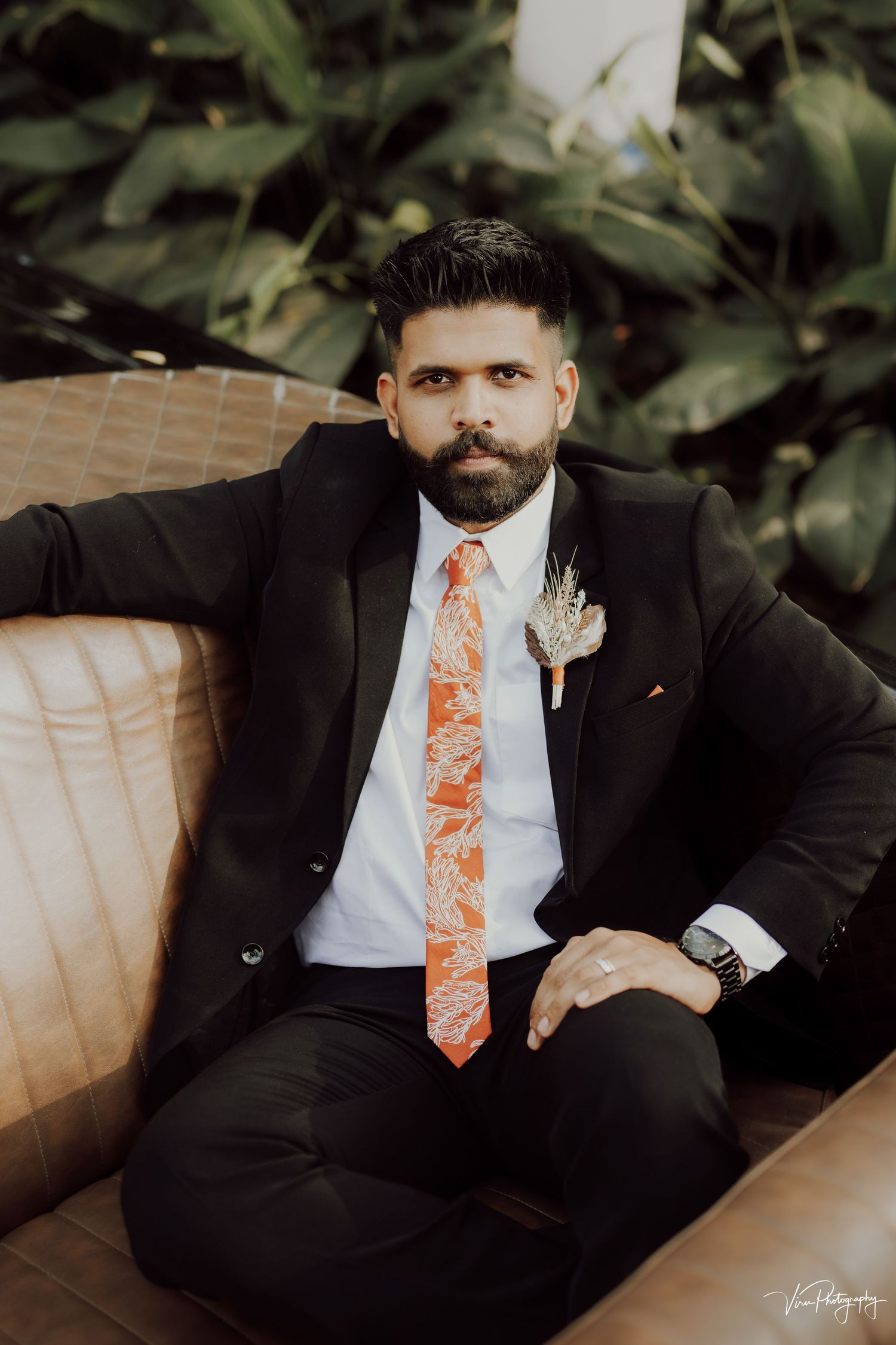 Groom in black suit with coral tie and boutonniere sits on tan couch during outdoor wedding photo session.