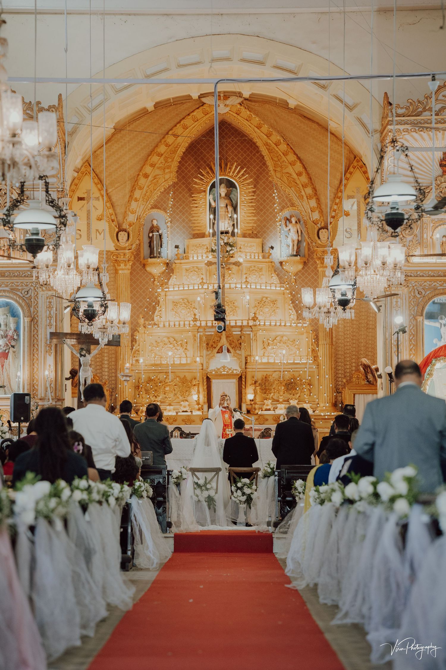 Interior view of an ornate Catholic church during a wedding ceremony with white decorations and a red carpet aisle.