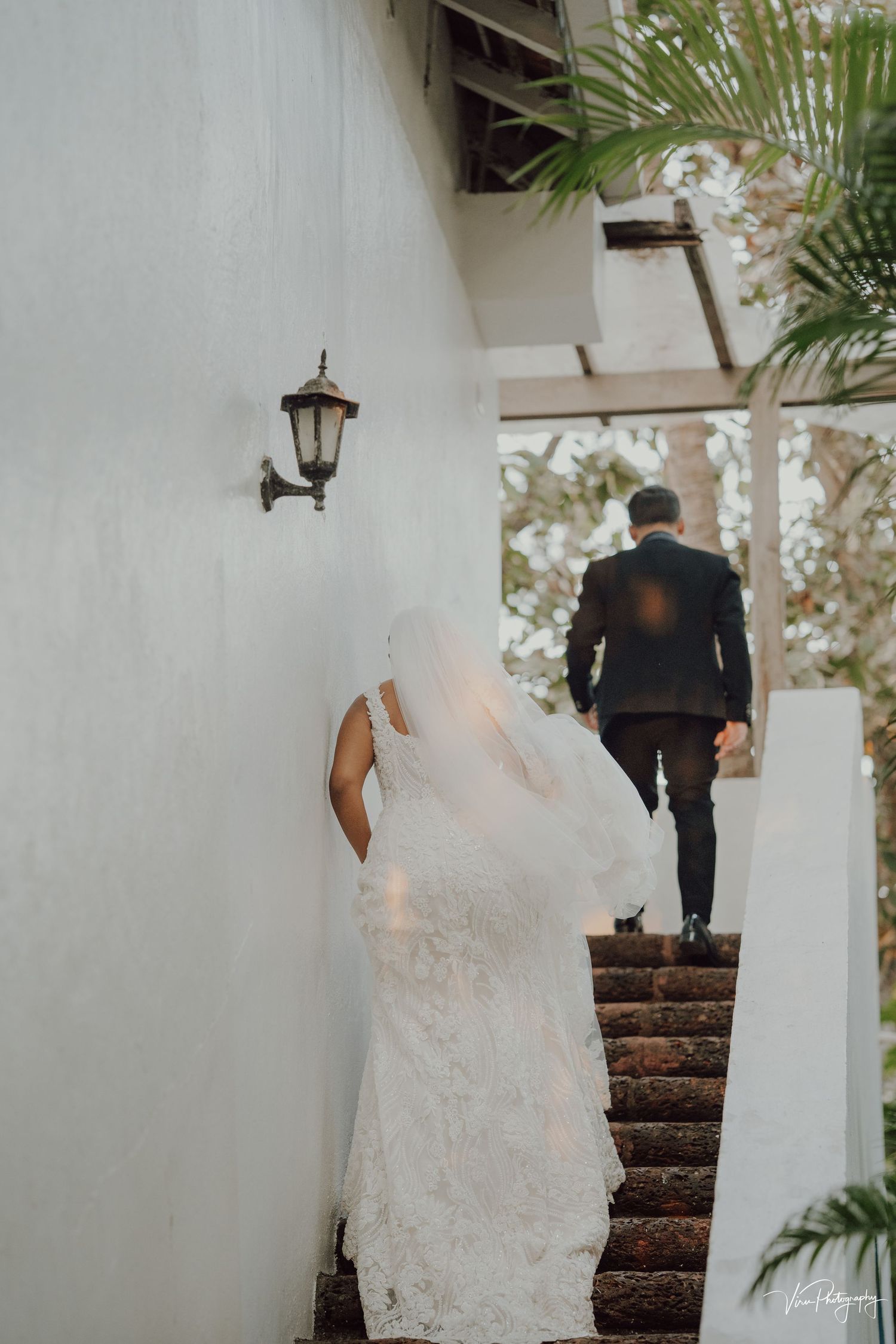 A bride in a lace white dress and groom in black suit ascend rustic stone stairs with a wall-mounted lantern.