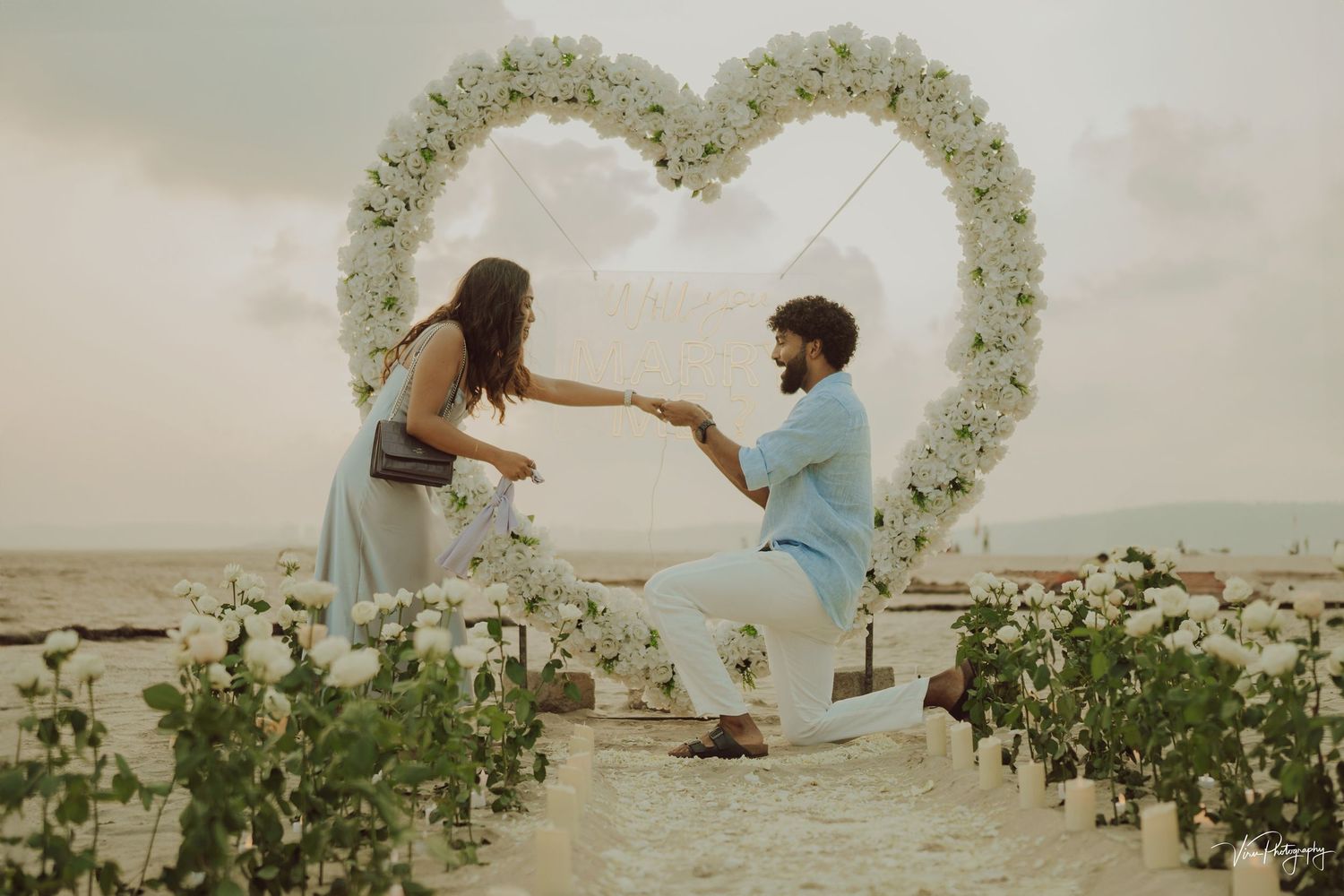 A romantic proposal scene under a floral heart-shaped arch on a cloudy beach with windswept greenery.