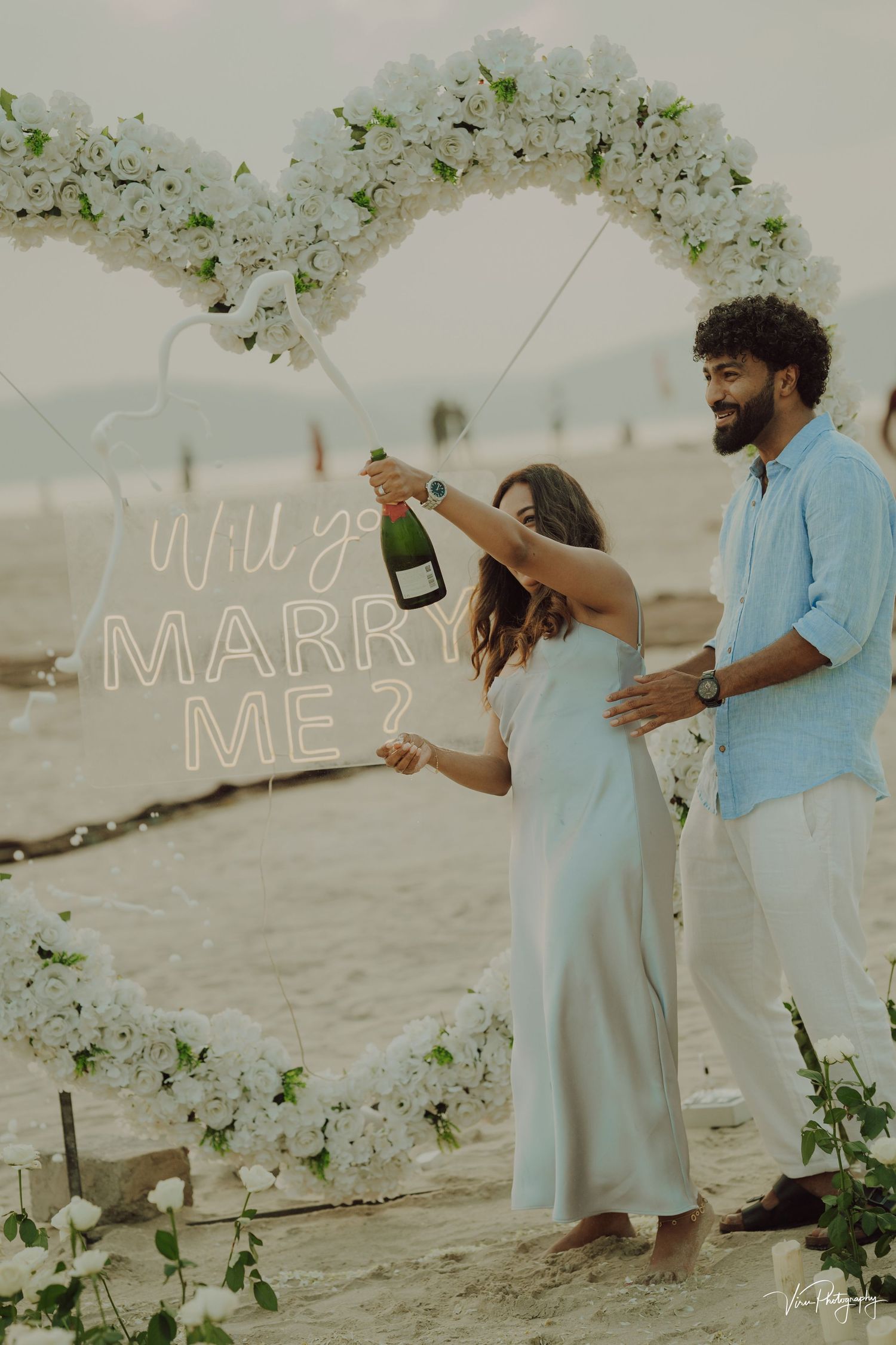 A romantic beach proposal setup featuring a white floral arch, neon sign, and champagne celebration at sunset.