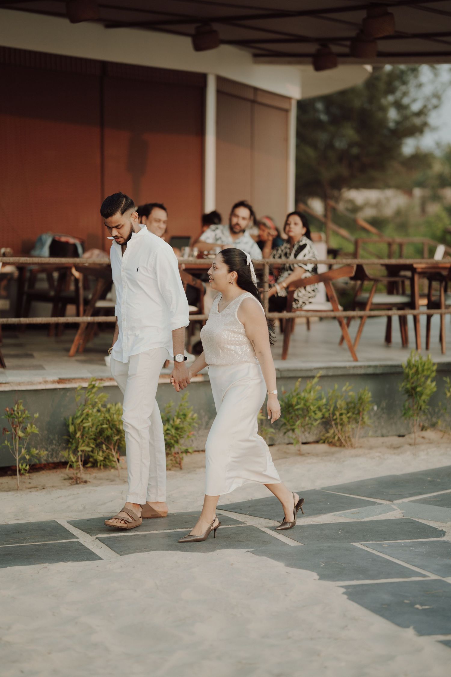Couple in white outfits walking hand in hand at an outdoor venue with guests seated in the background.