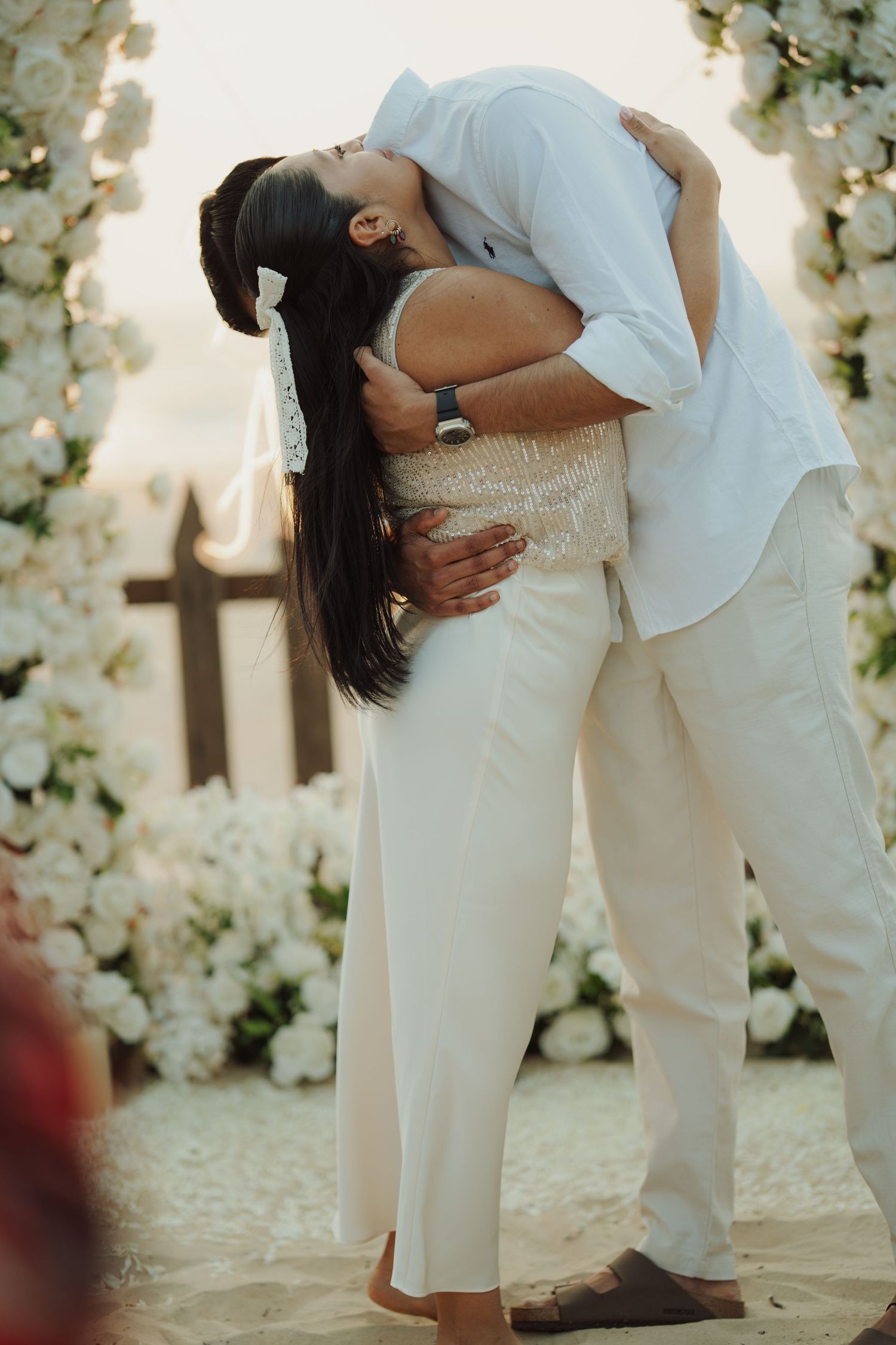 Couple sharing a romantic kiss at a beach wedding ceremony surrounded by white floral arrangements.