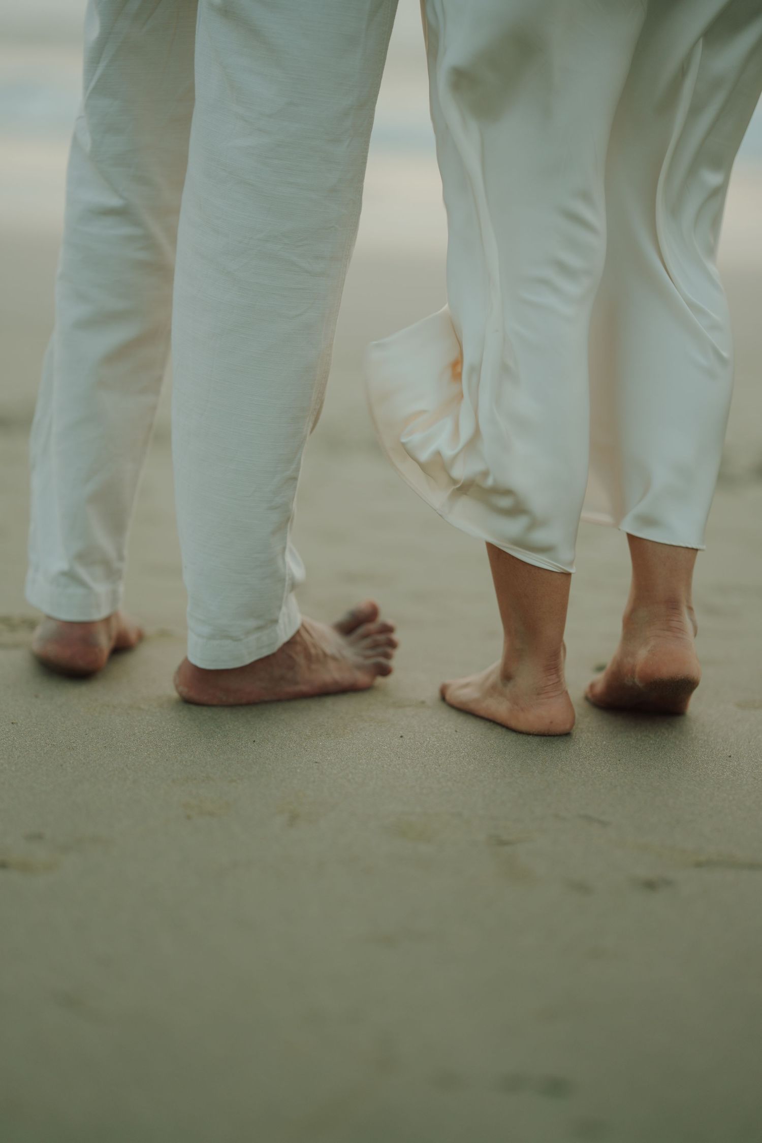 Barefoot couple in white linen pants standing on sandy beach, close together, soft natural light.
