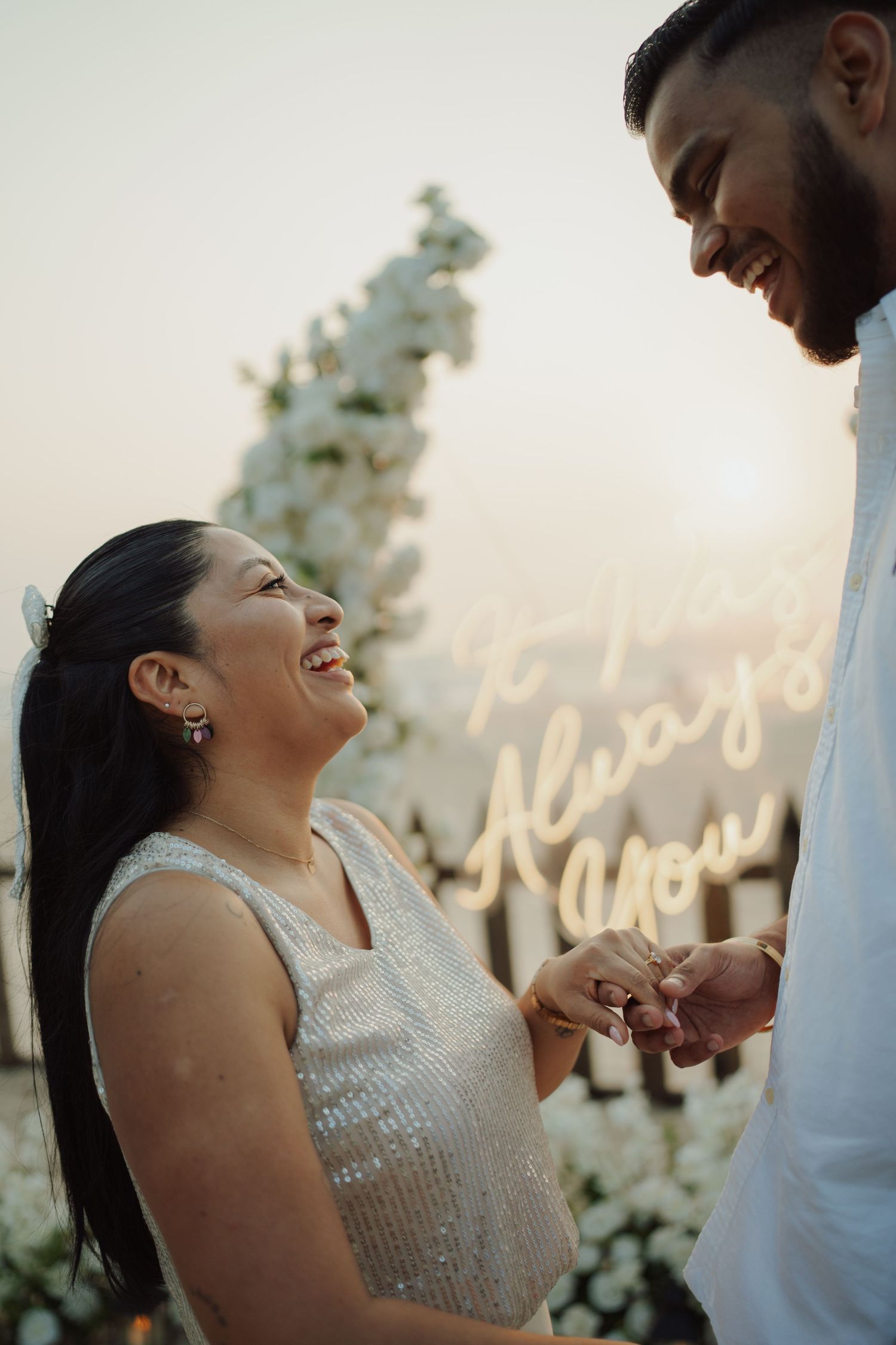 Joyful bride laughing as groom places ring on her finger at sunset beach proposal with neon sign.