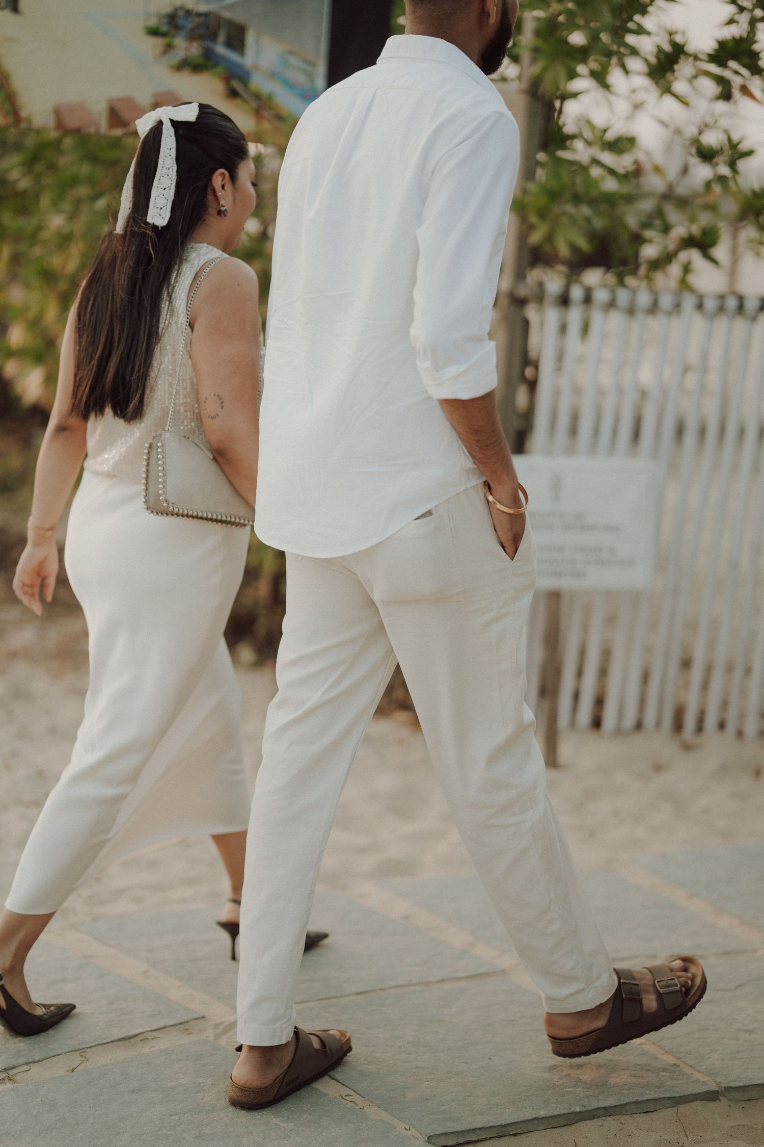 Couple walking in matching white outfits, woman with bow hair accessory, man in linen shirt and pants.