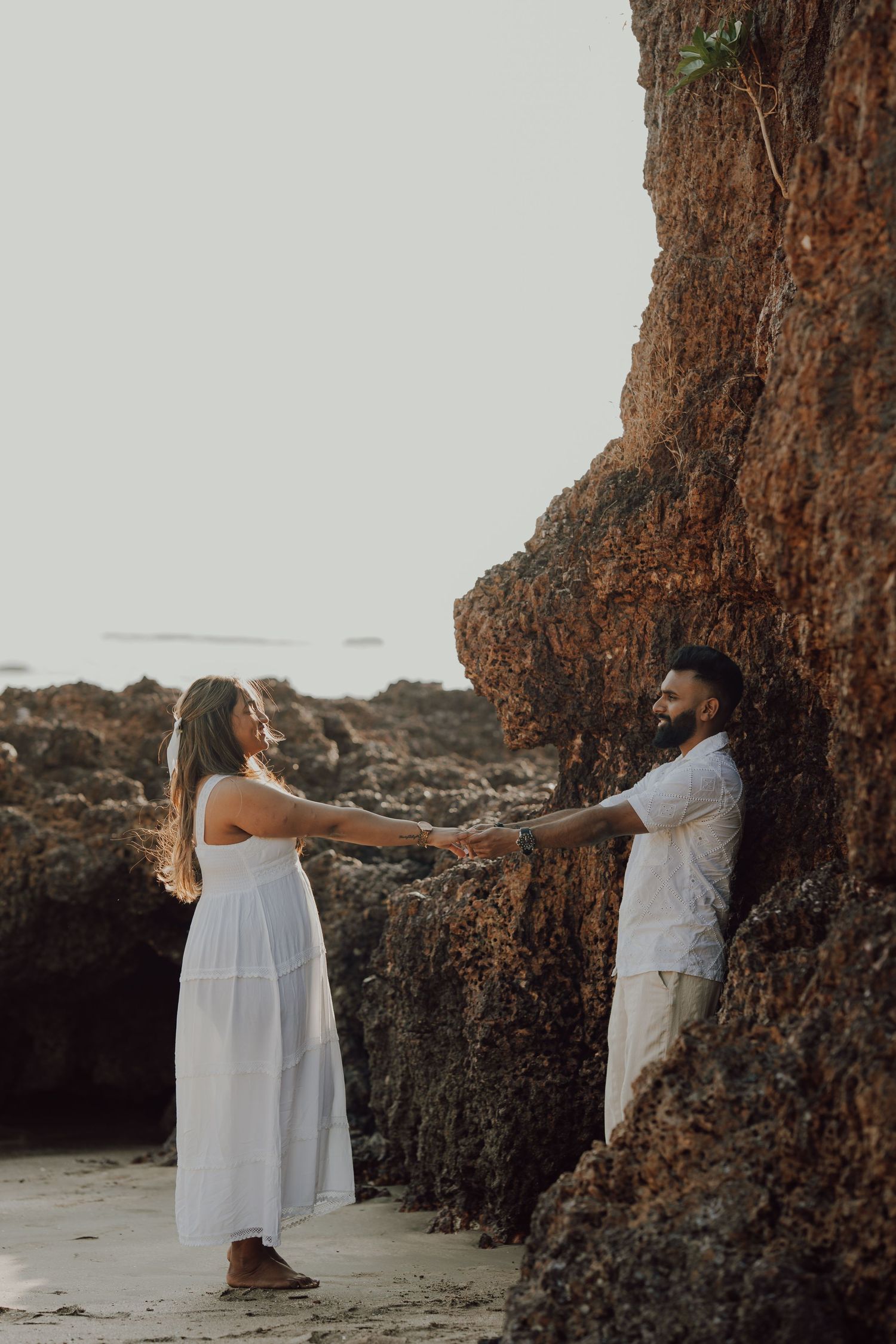 A couple in white outfits embraces against rocky cliffs at sunset on a beach, creating a romantic silhouette.