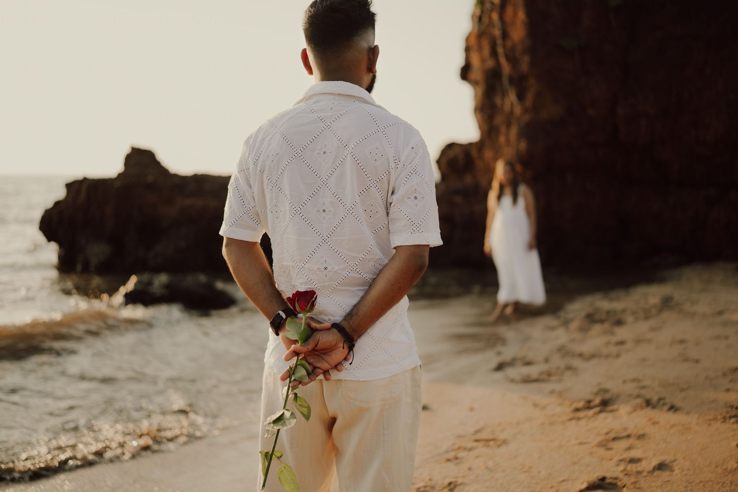 Couple in white attire sharing an intimate moment on a rocky beach at sunset.