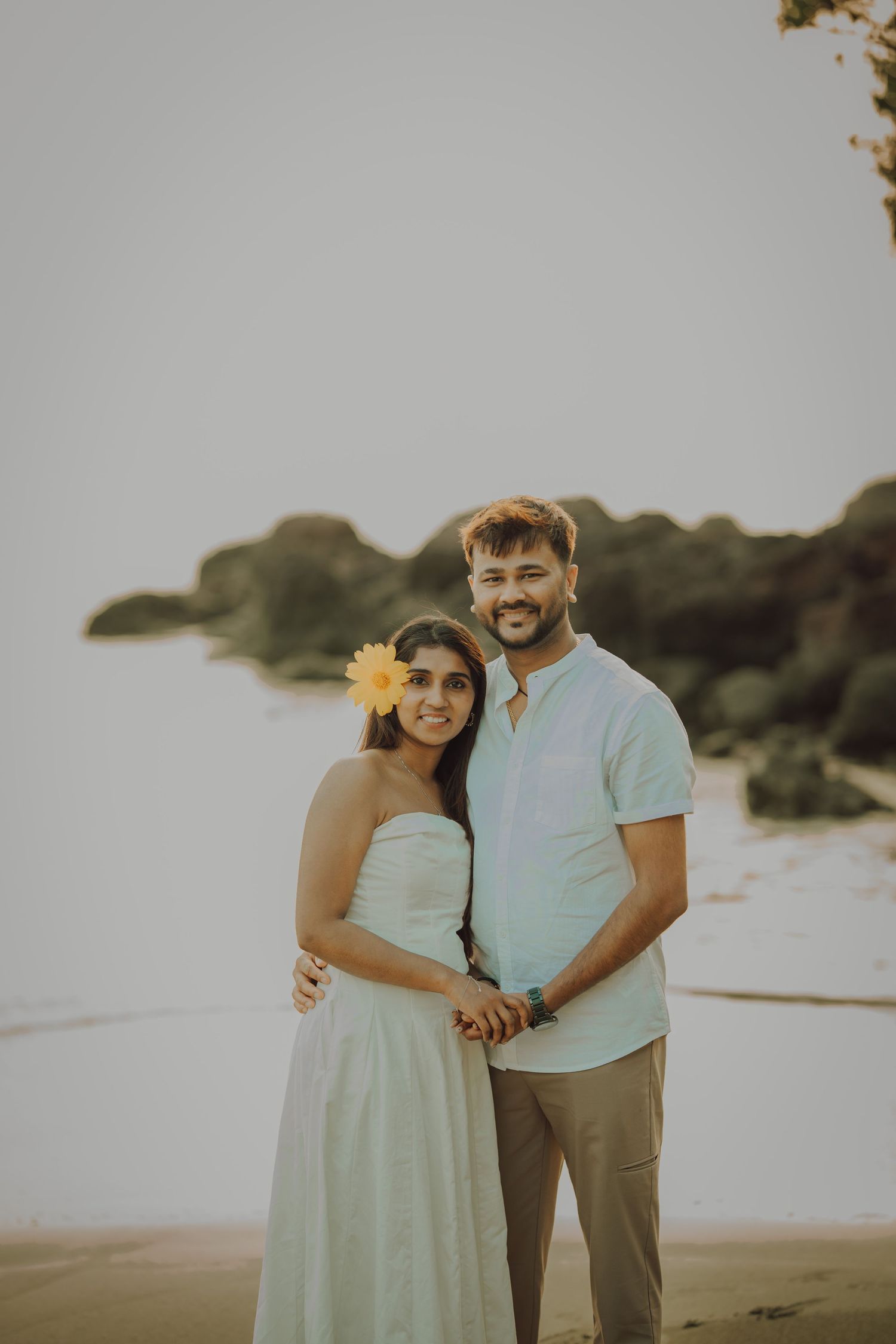A couple stands together on a beach at sunset wearing light colored clothing and sharing an intimate moment.