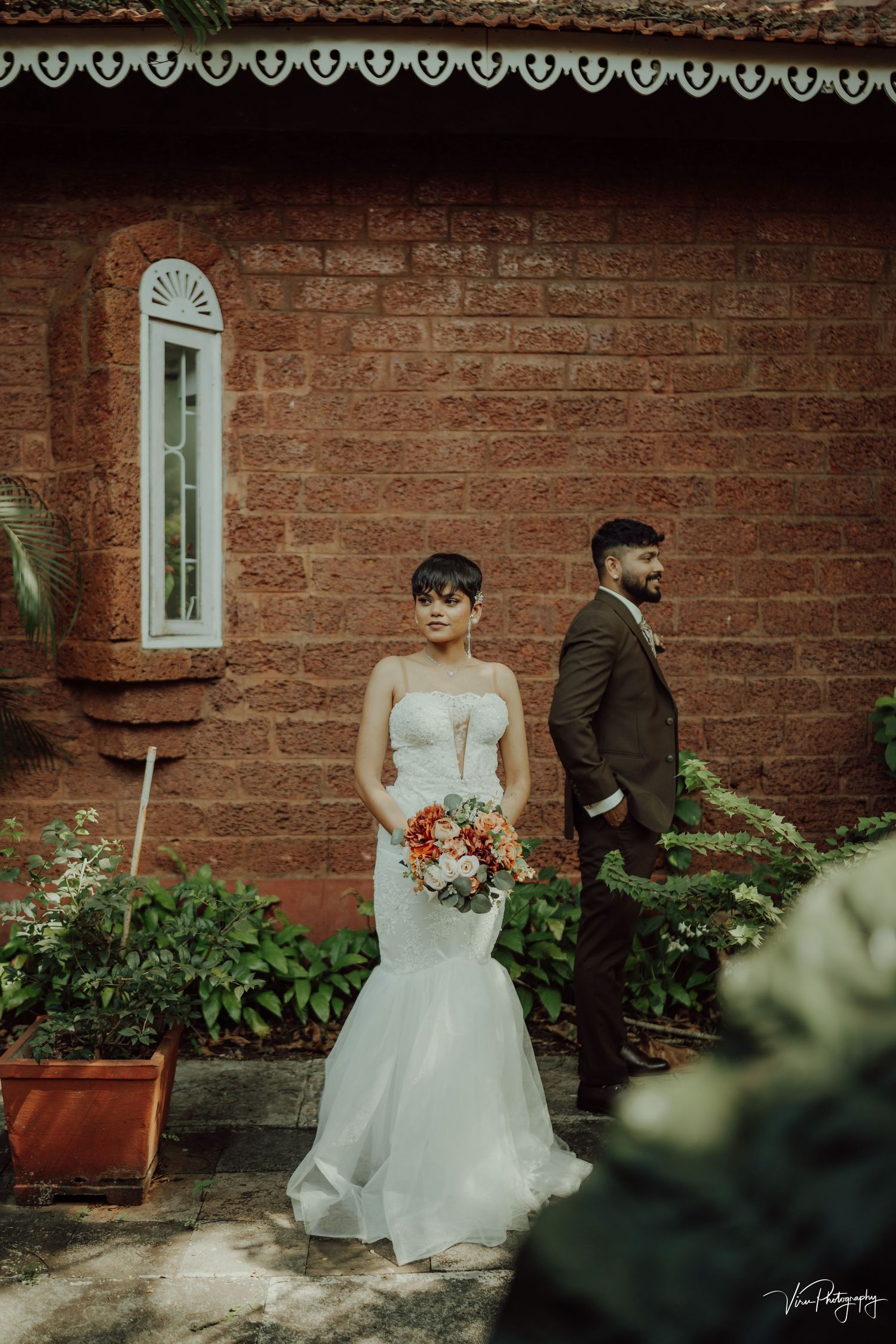 A couple poses separately against a rustic brick wall with vintage window details and potted garden plants.
