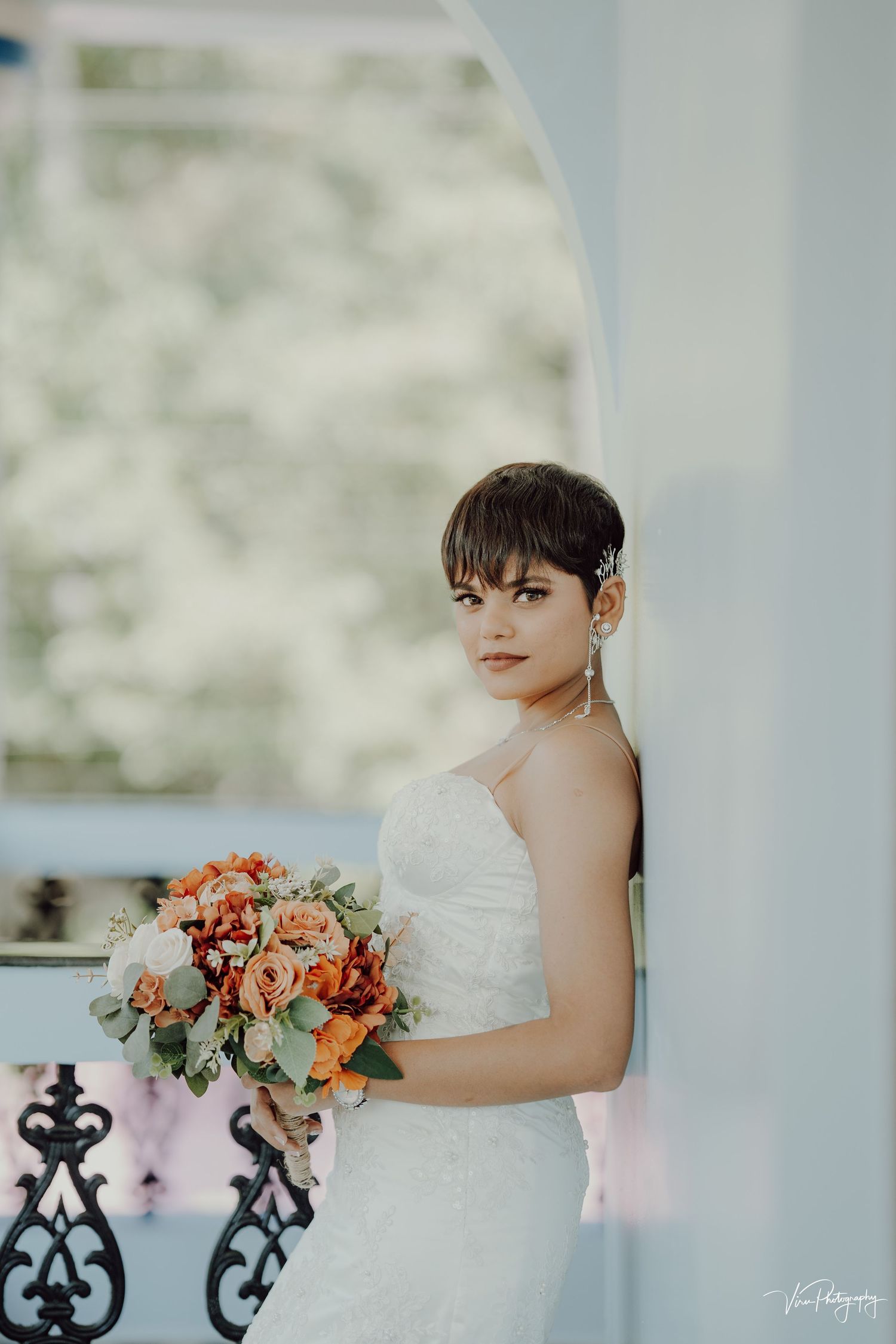 A bride in a white wedding dress poses elegantly next to a decorative table with orange and pink floral arrangements.