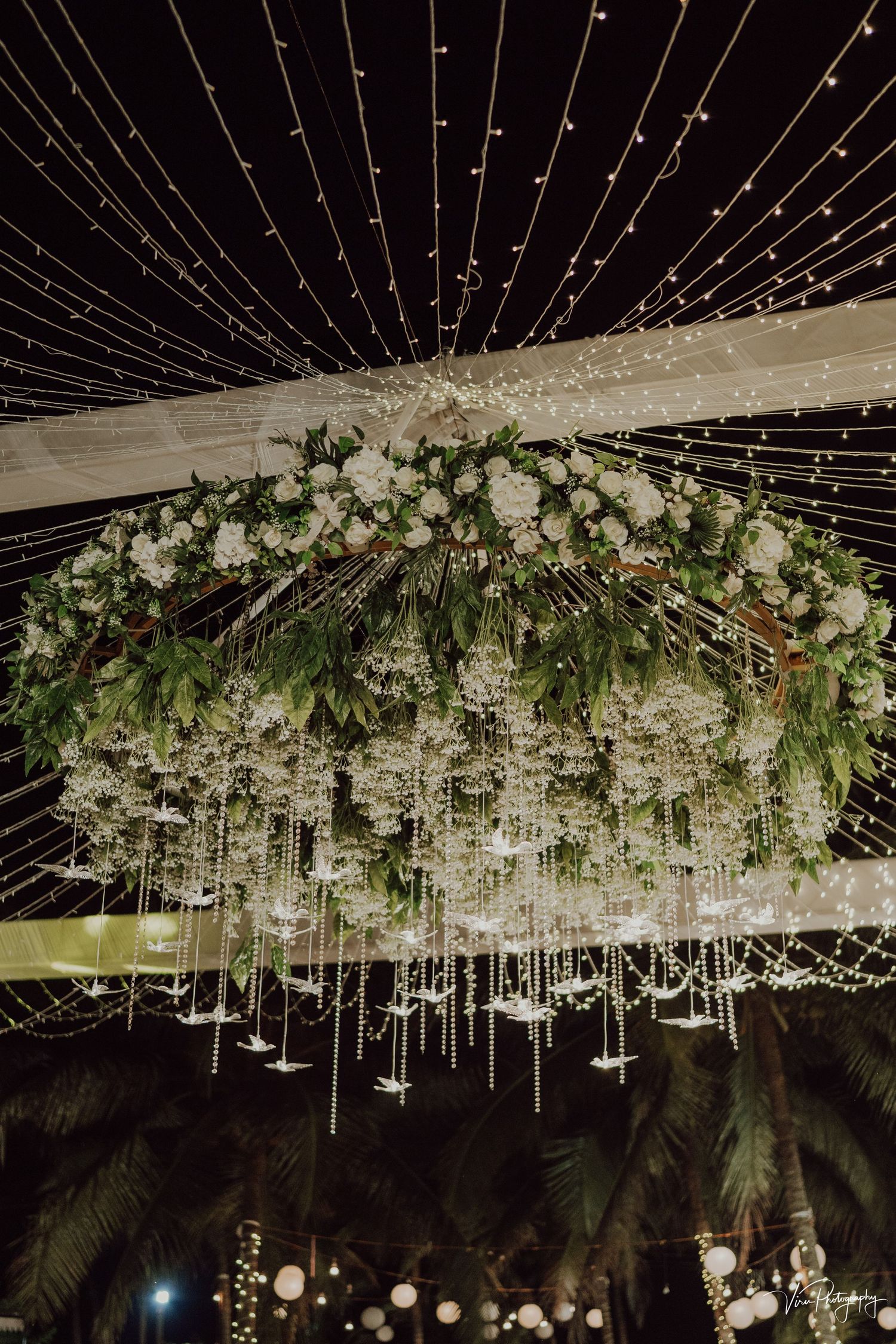 Elegant wedding reception chandelier decorated with white flowers, hanging crystals and twinkling string lights against night sky.