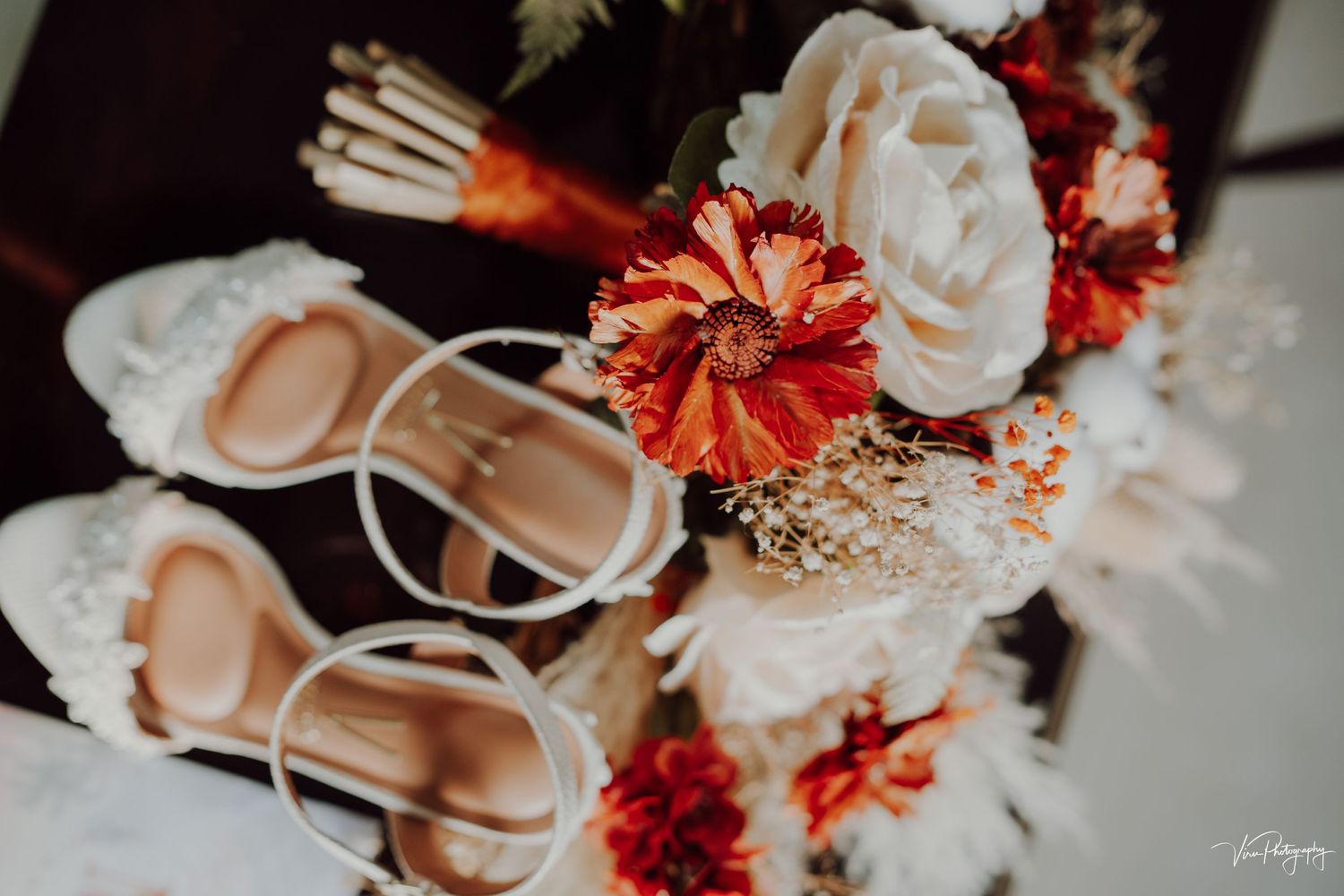 White wedding shoes and a bridal bouquet with white roses and orange flowers arranged on light surface.