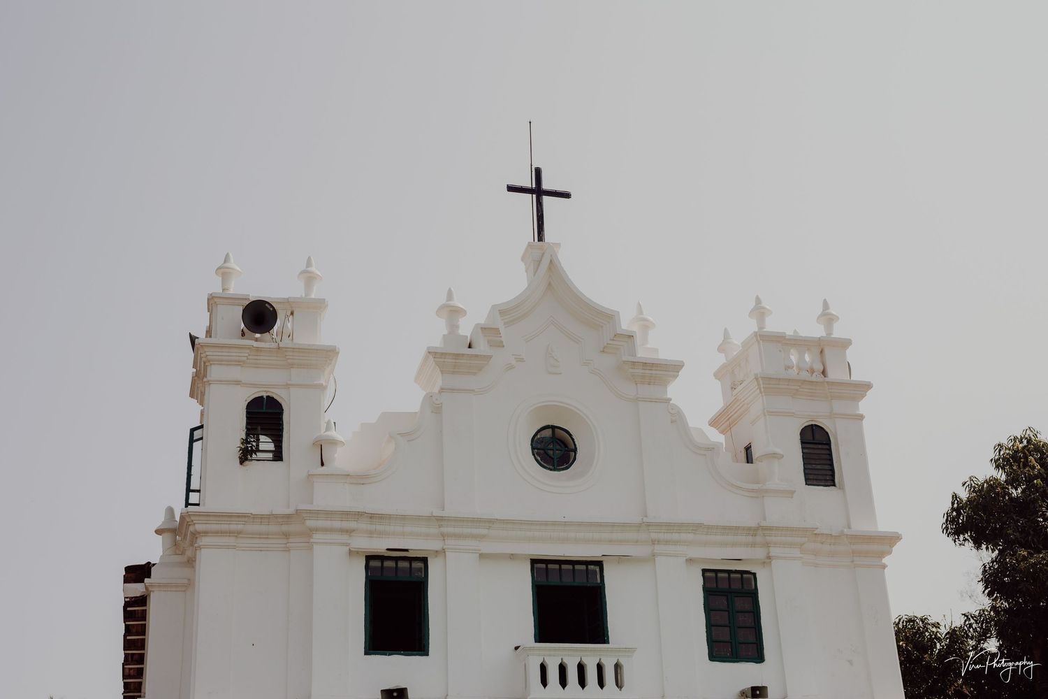 A white Portuguese colonial church with three bell towers and cross stands against an overcast sky at sunset.