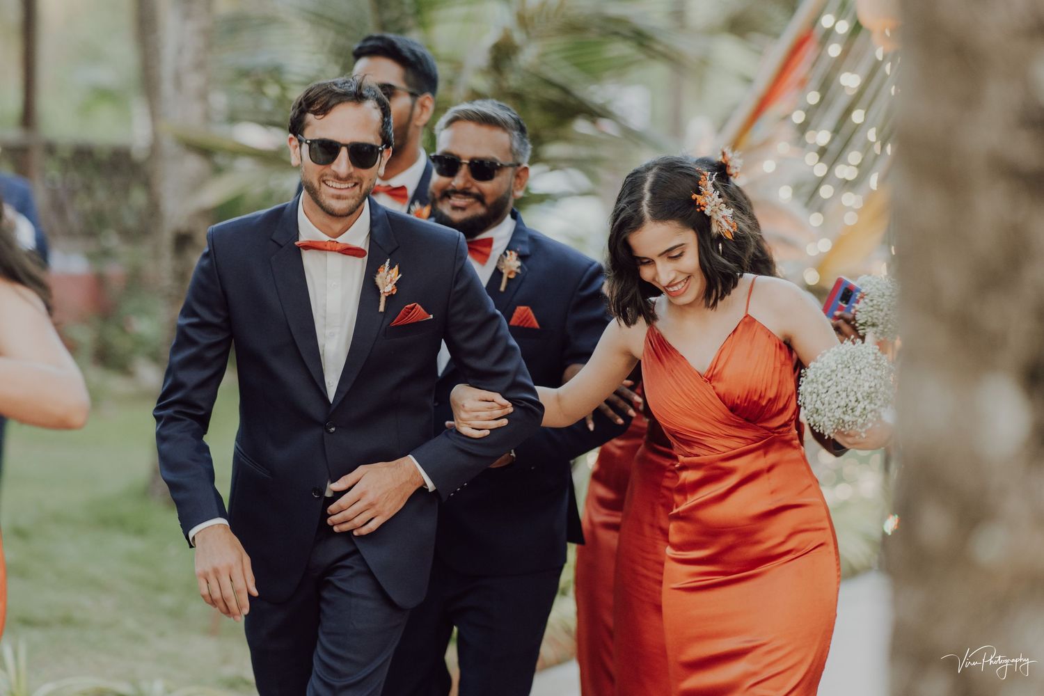 A person in an orange dress walks arm-in-arm with two others wearing dark suits at an outdoor event.