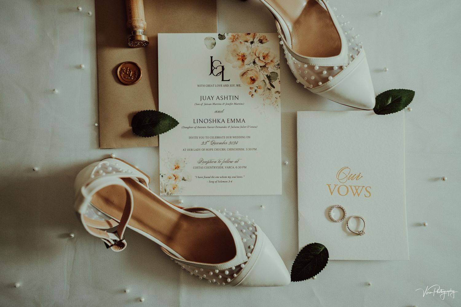 Elegant white bridal shoes with floral details arranged next to wedding invitation and gold rings on light background.