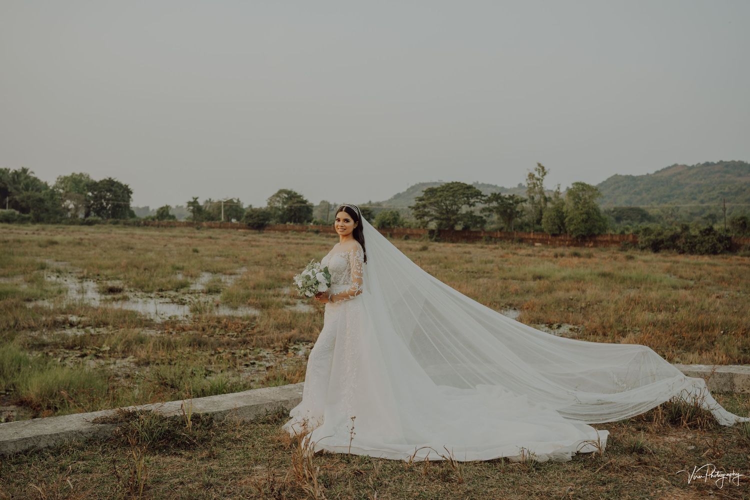 A person in an elegant white wedding dress with flowing veil stands in a serene meadow with mountains in the background.