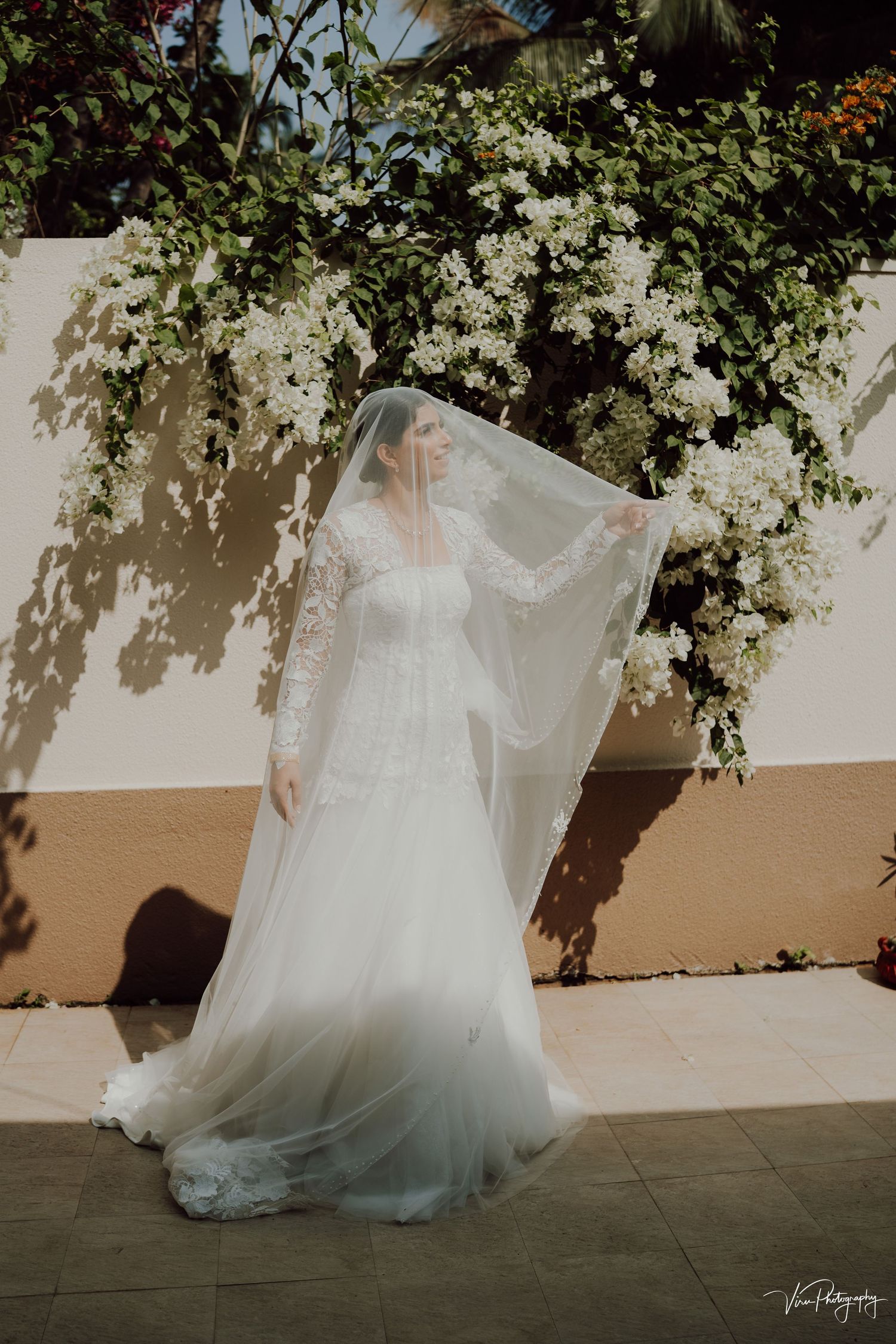Elegant white wedding gown and veil draped against a wall covered in cascading white bougainvillea flowers in sunlight.