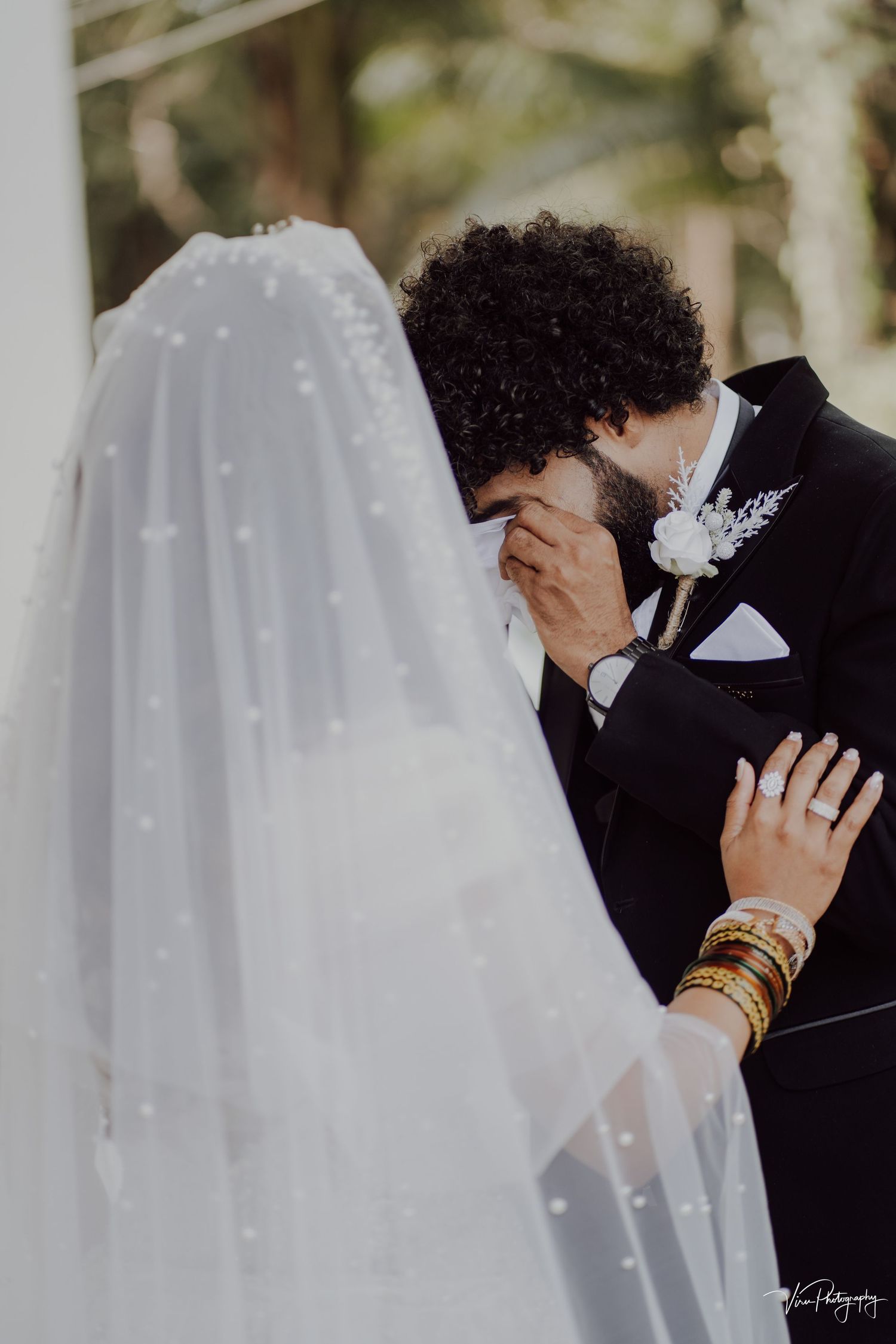 An intimate wedding moment as a couple embraces, with the bride in a pearl-dotted veil and groom wearing a floral boutonniere.