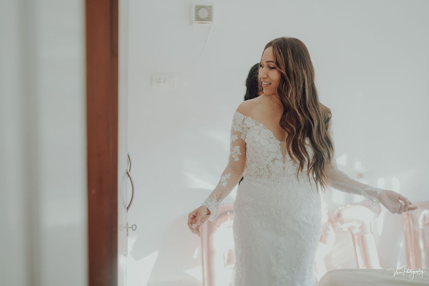 A bride in a lace wedding dress with flowing sleeves stands elegantly in a brightly lit room.
