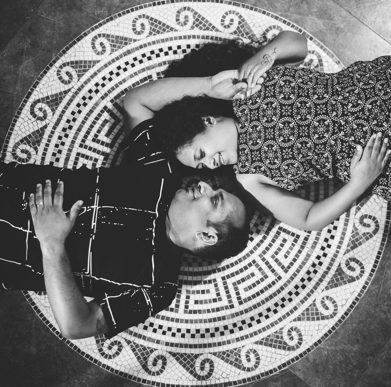 Black and white artistic overhead photo of two people lying on a patterned circular rug in dramatic composition.