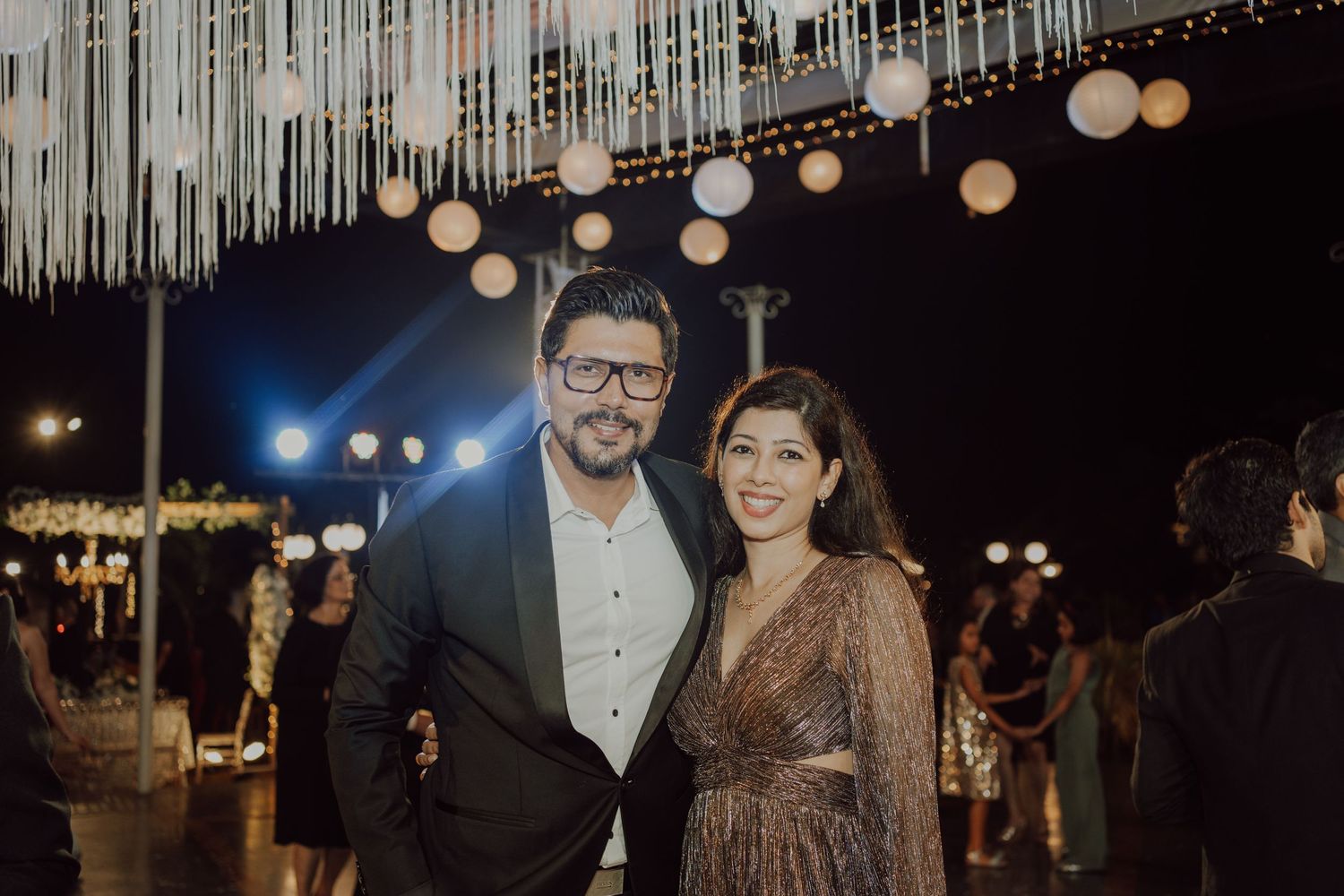 Elegantly dressed couple poses at evening event under decorative string lights.
