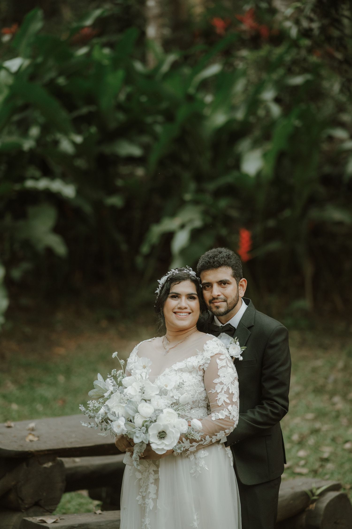 A wedding couple poses together in an outdoor garden setting with white floral bouquet.