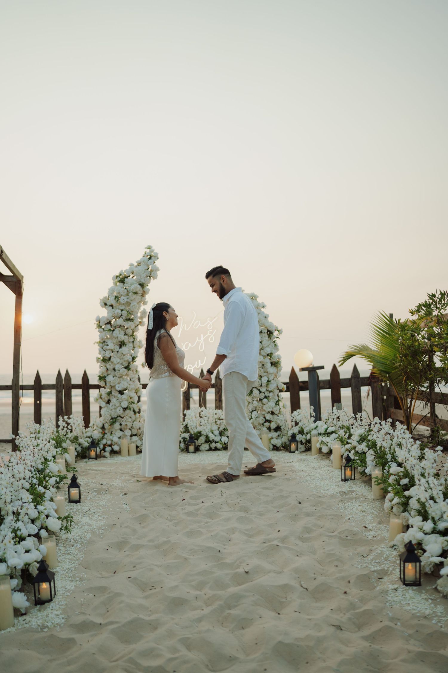 Couple holding hands at a romantic beach wedding ceremony with white floral arch and lanterns at sunset.