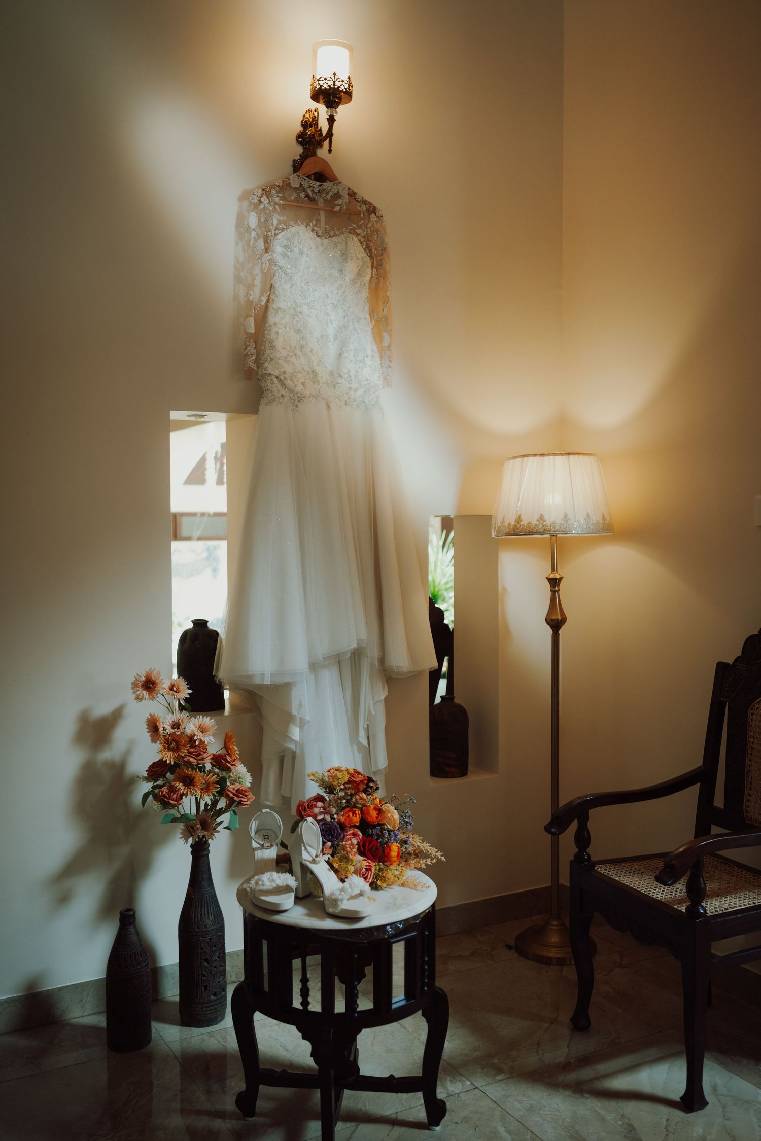A lace wedding dress hangs from a wall sconce above a small table with bridal shoes, a colorful flower bouquet, vases, and a floor lamp.