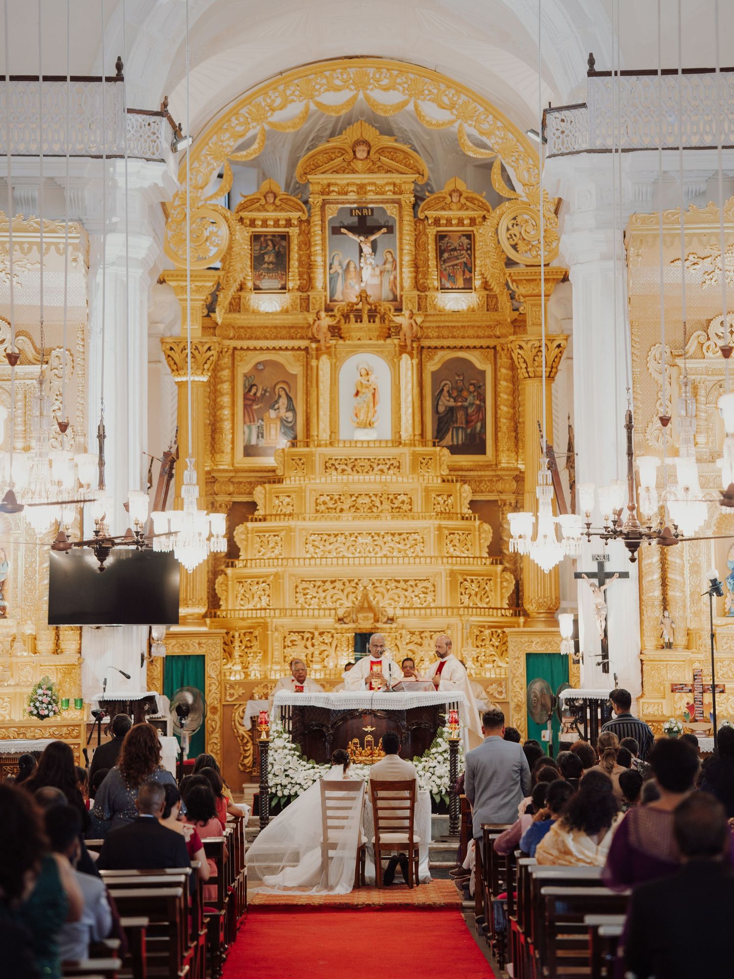 Wedding guests watch the ceremony inside an ornate church with a golden baroque altar and a red carpet aisle.