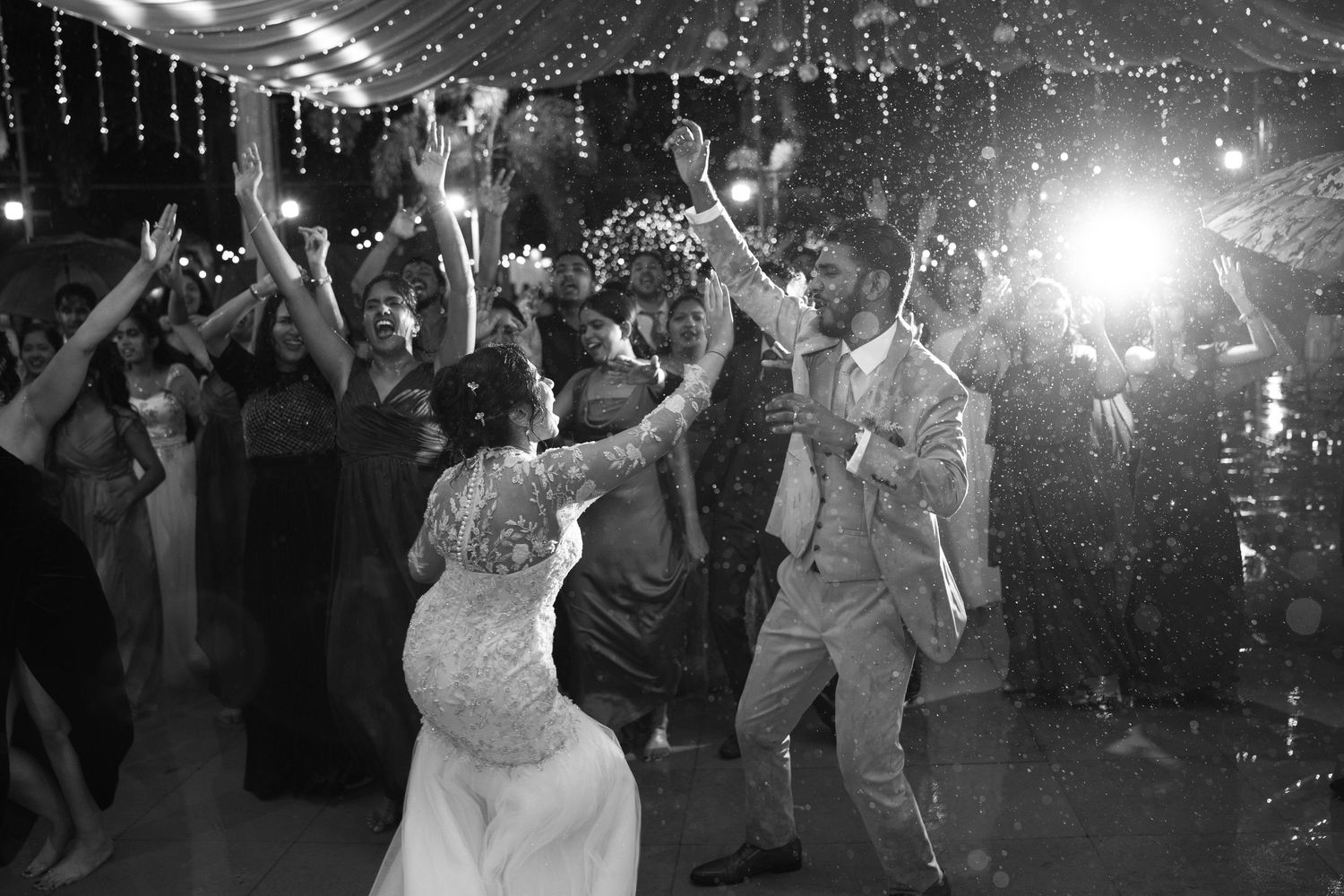 Black and white photo of a bride and groom dancing joyfully with cheering wedding guests under bright lights at the reception.