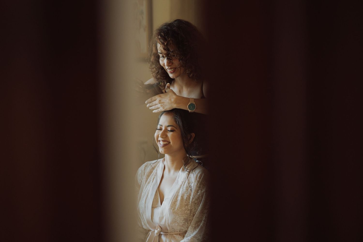 Smiling woman in a white robe having her hair styled by a curly-haired woman, framed by dark curtains in warm natural light.