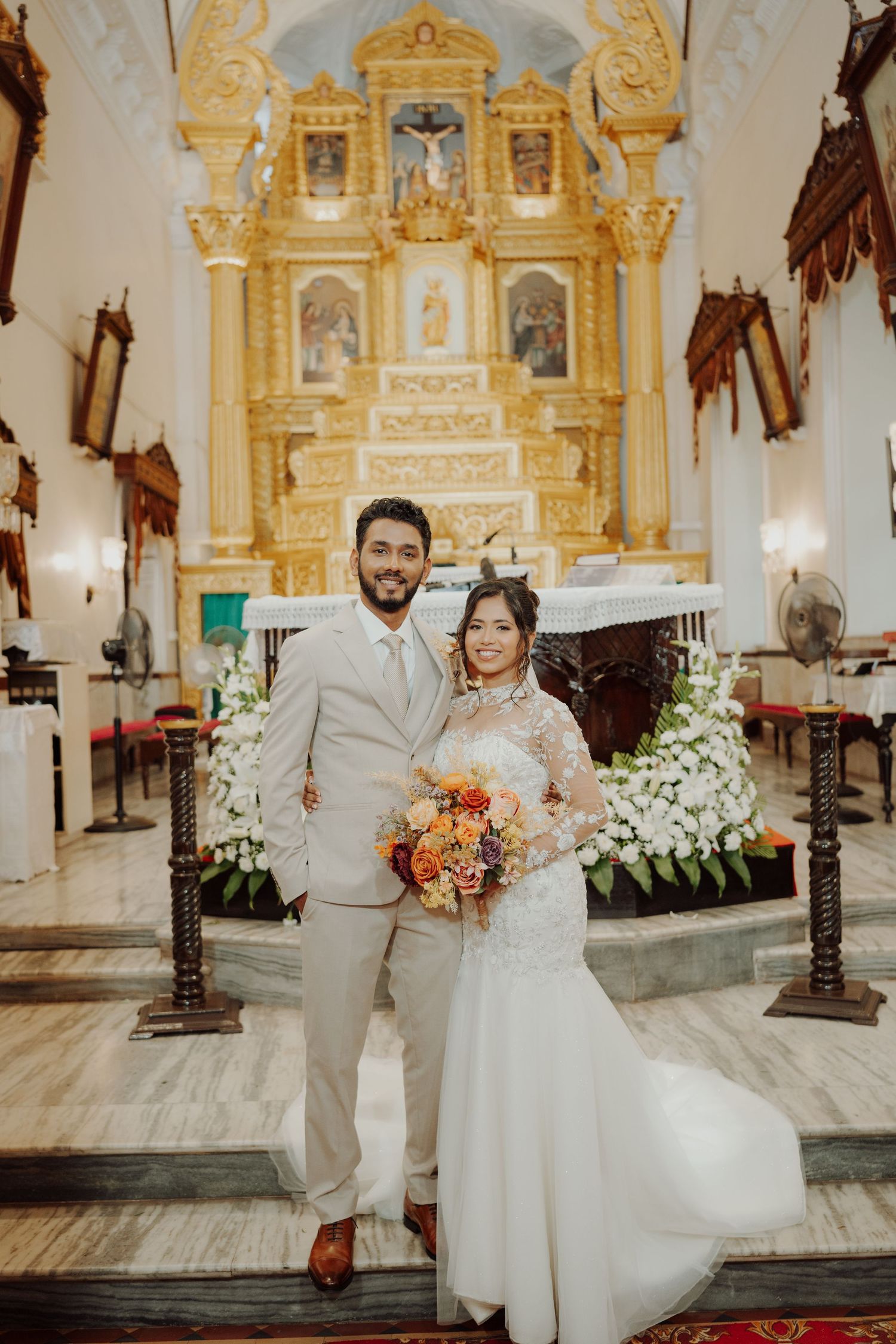 A smiling bride and groom in a beige suit pose with an orange bouquet before a golden church altar adorned with white flowers.
