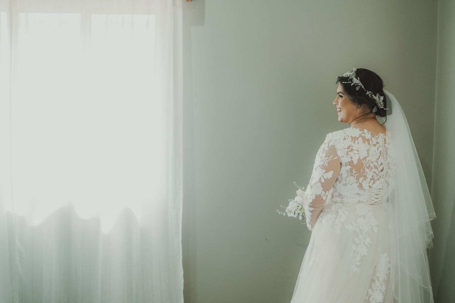 A bride in a lace wedding gown stands by a window in soft natural light.