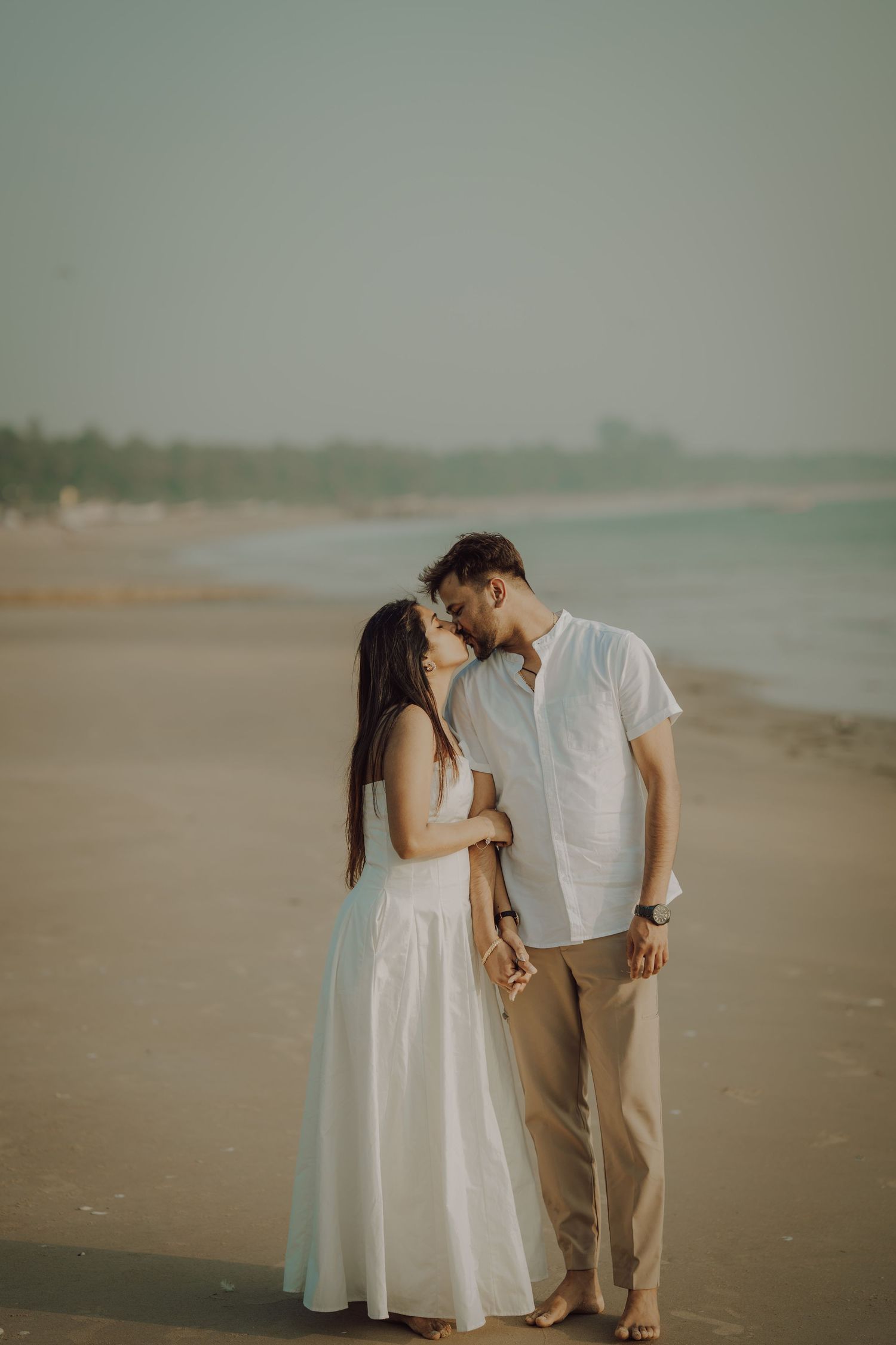 A romantic couple shares intimate moments on a misty beach while wearing white and beige outfits.