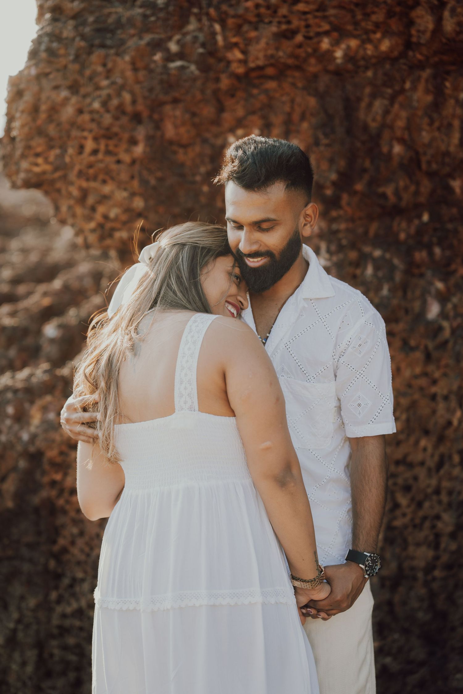 A romantic couple in white outfits share intimate moments against a rustic stone wall background.
