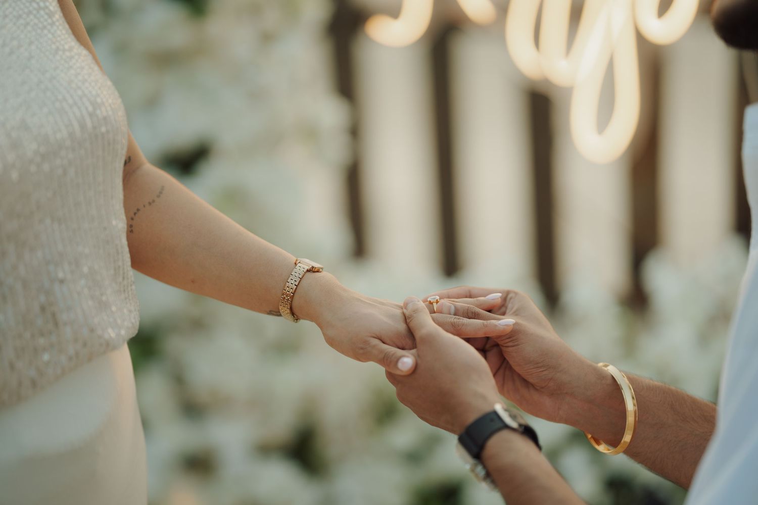 Couple exchanging rings at wedding ceremony with gold bracelets and bokeh floral backdrop.