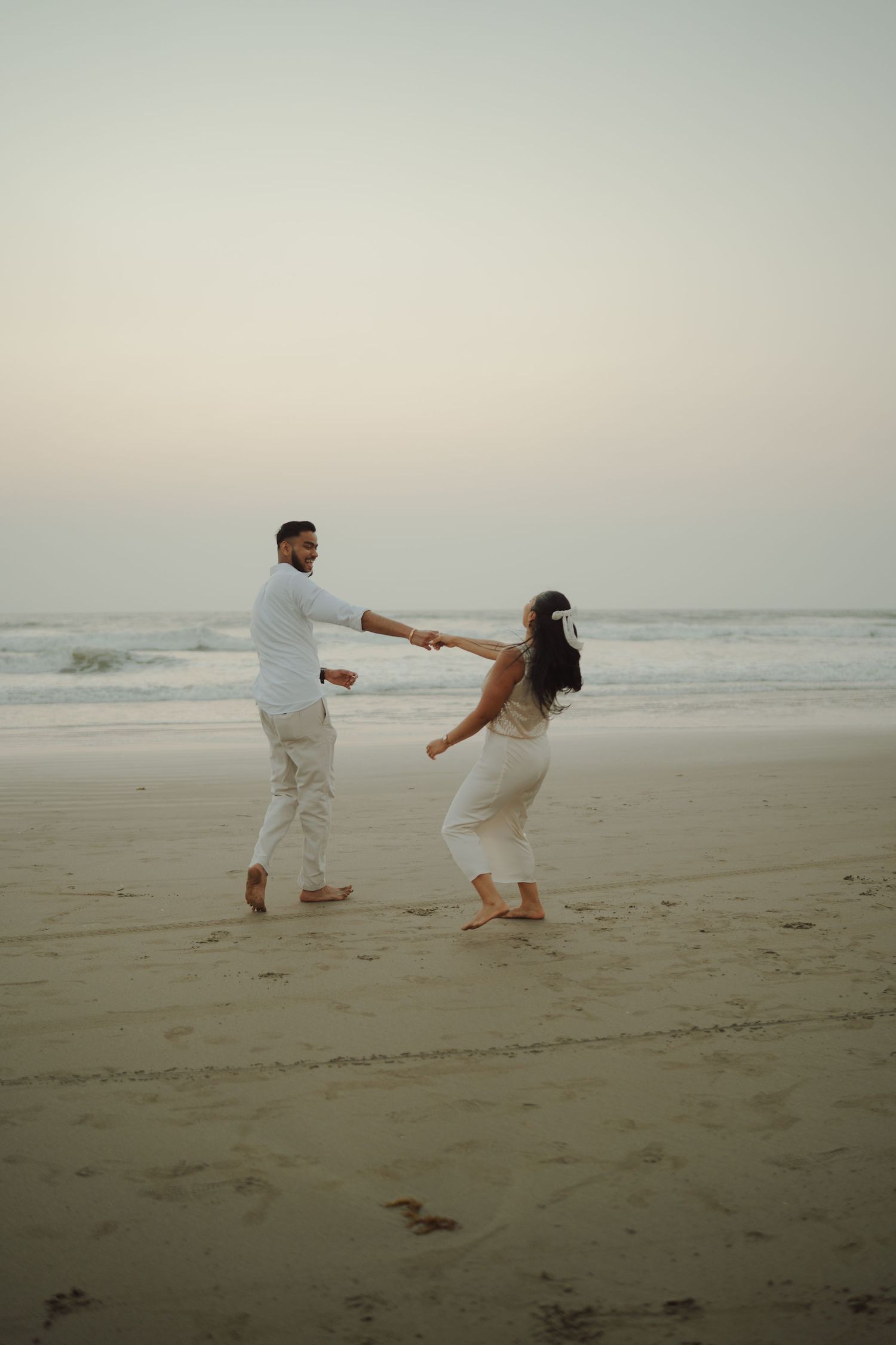 Couple in white outfits dancing barefoot on a sandy beach at dusk, holding hands near ocean waves.