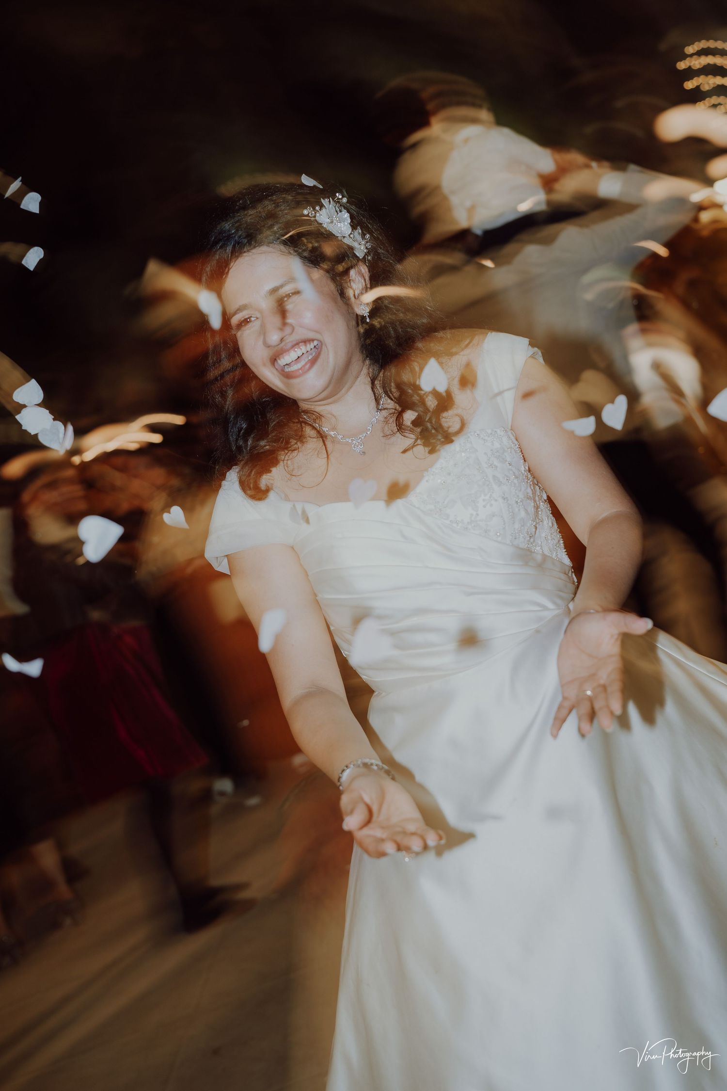 A joyful bride in a flowing white dress dances during her wedding reception as flower petals float through the air around her.