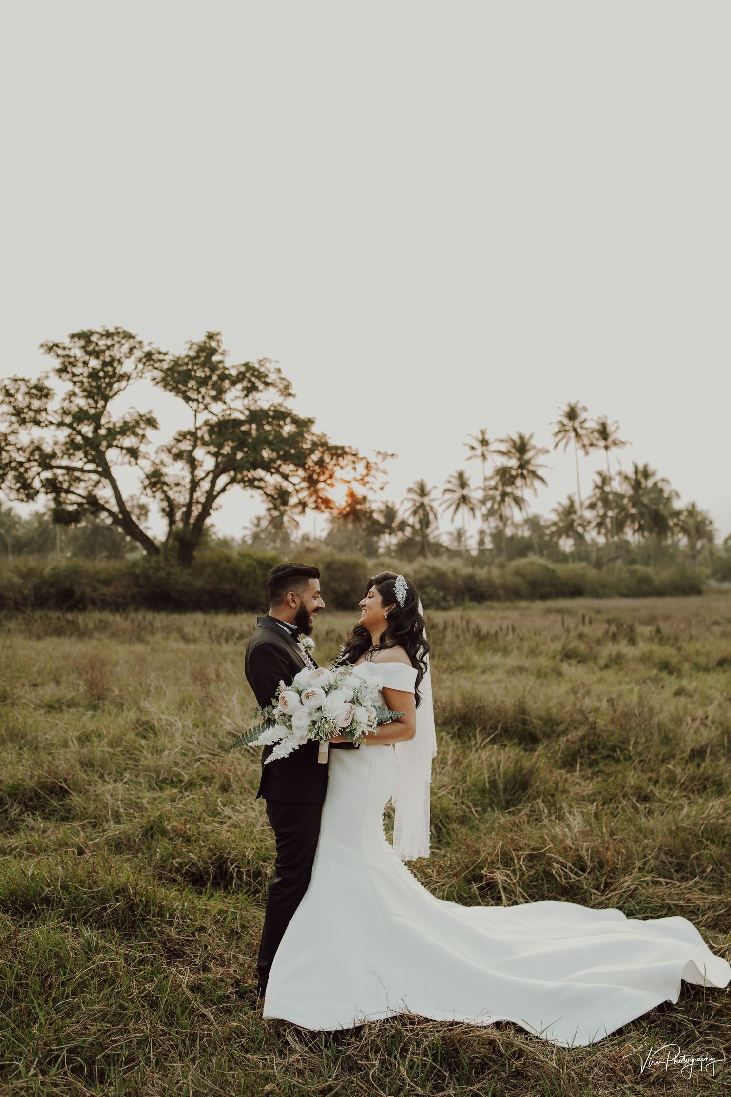 A romantic wedding couple embraces in an outdoor field at sunset with palm trees and flowing white dress in the background.