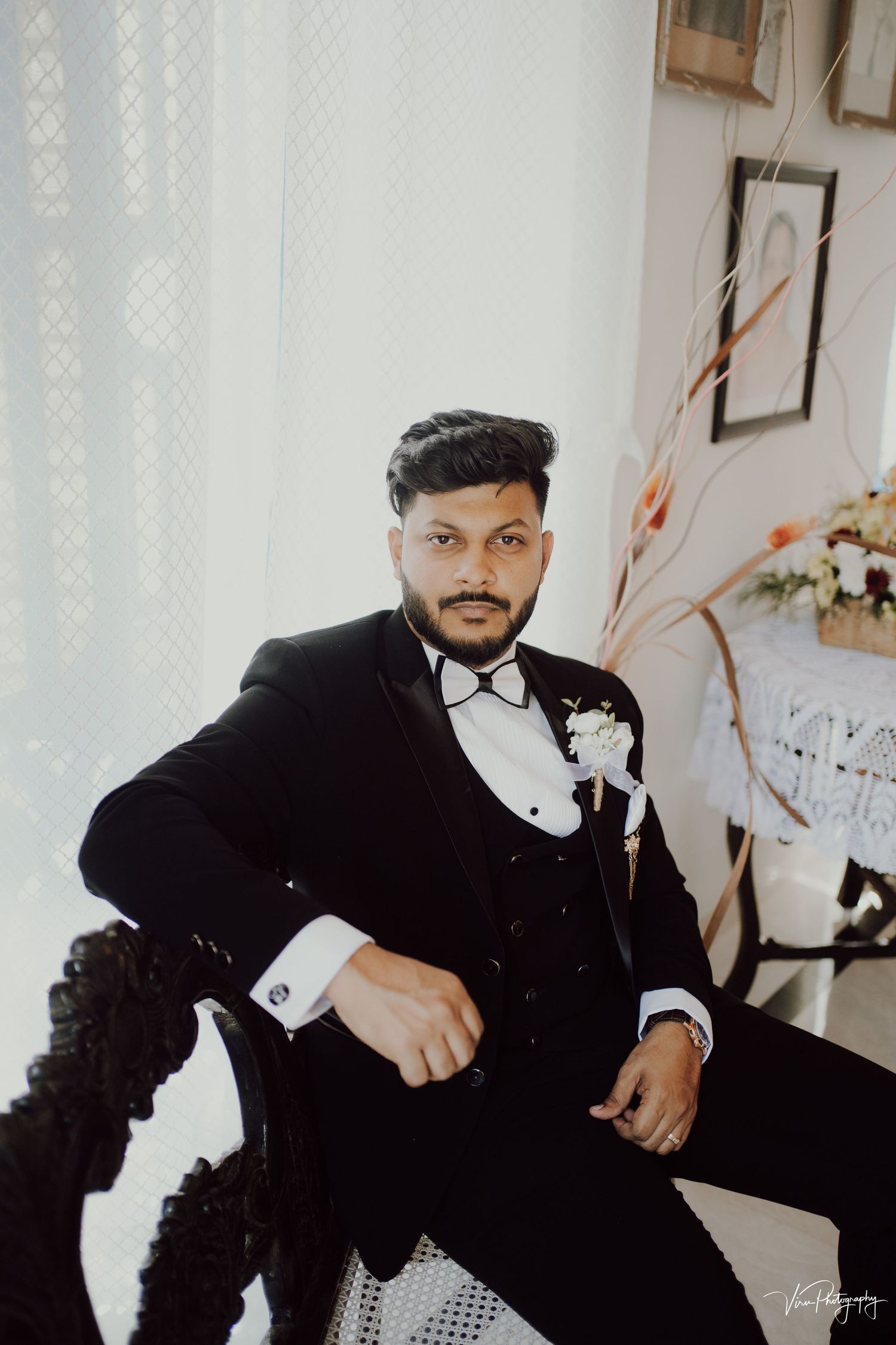 Sophisticated person in formal black tuxedo with bow tie and boutonniere poses elegantly in light-filled indoor setting.