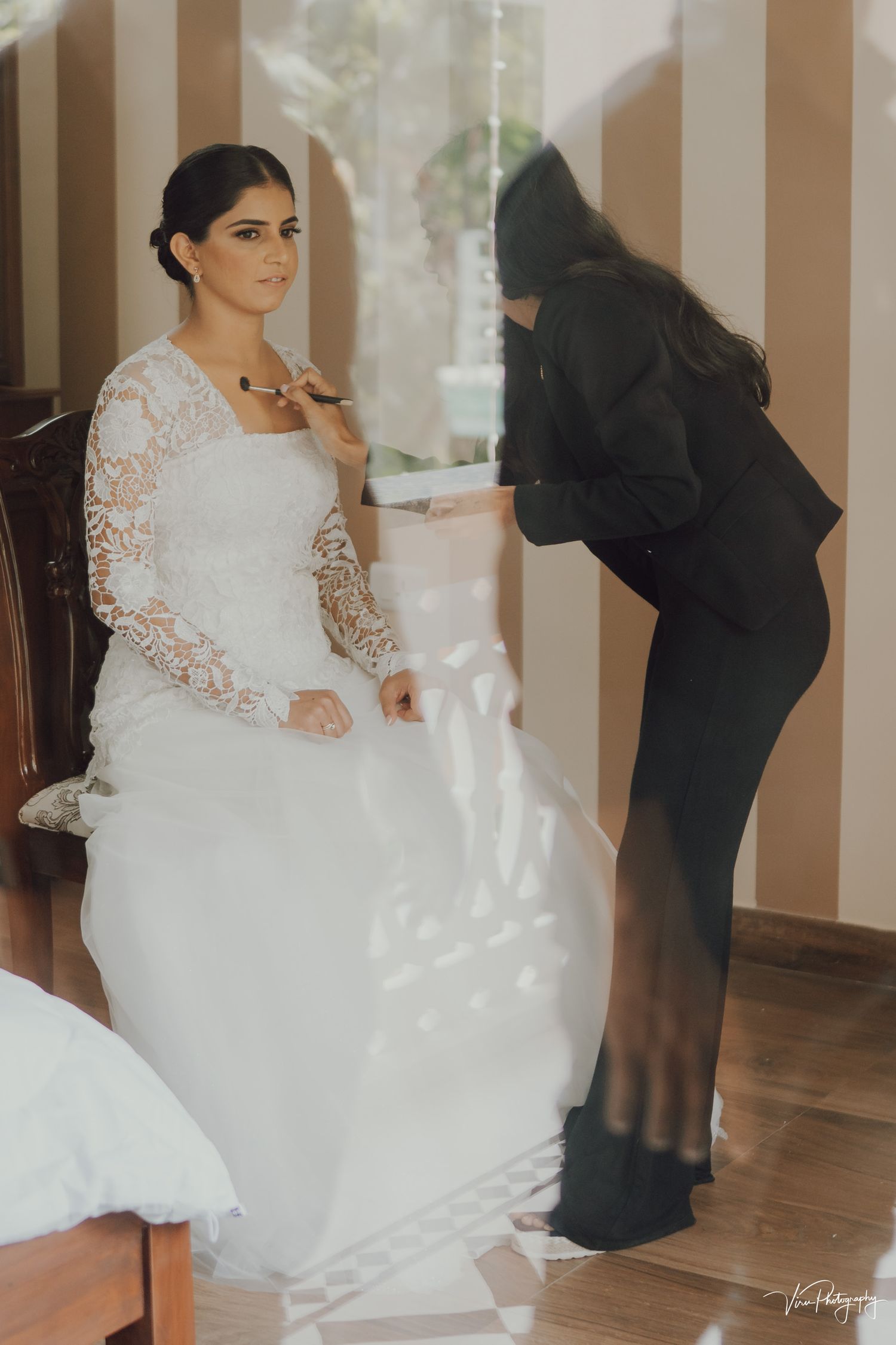 A person in a white lace long-sleeved wedding dress being assisted with final preparations in a light-filled room.