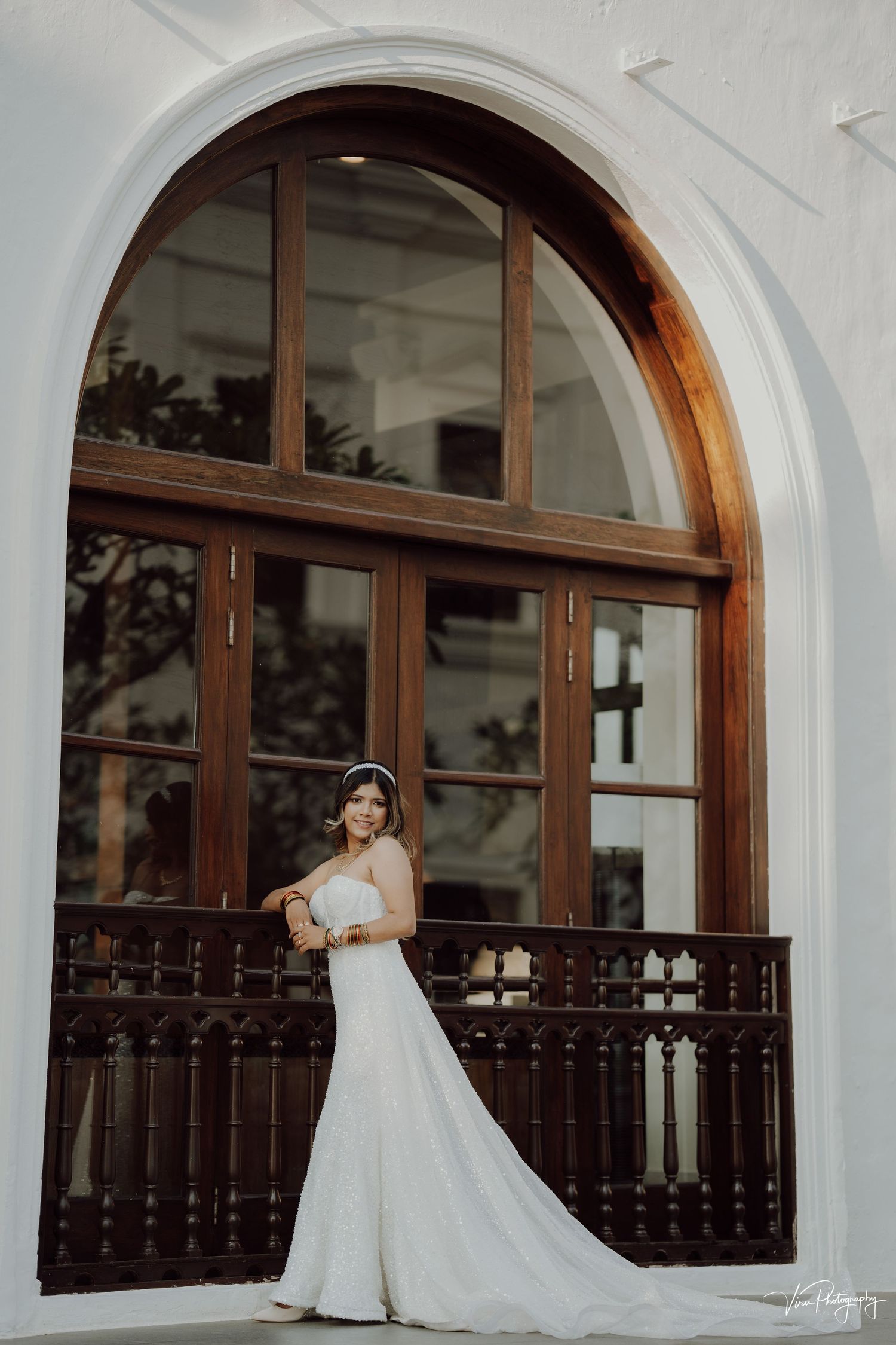 A bride in a flowing white wedding gown stands elegantly on a balcony framed by a grand arched wooden window.
