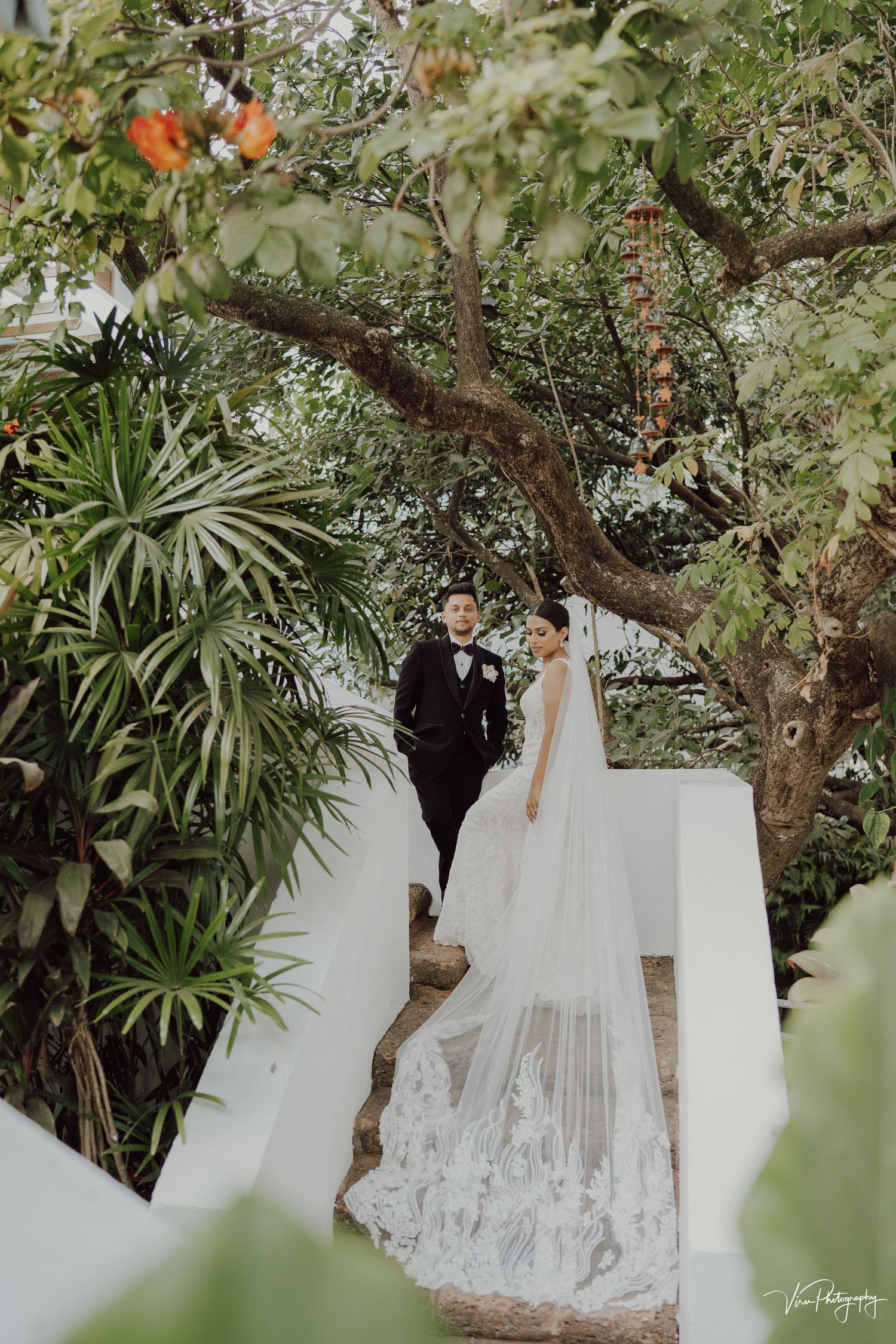 A couple poses in a lush tropical garden setting with a flowing white lace wedding dress veil.