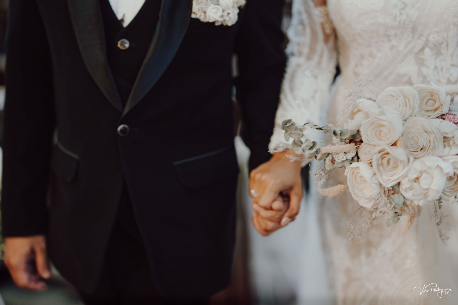 Close-up shot of a bride and groom holding hands, showing the details of their wedding attire and a blush-colored bouquet.