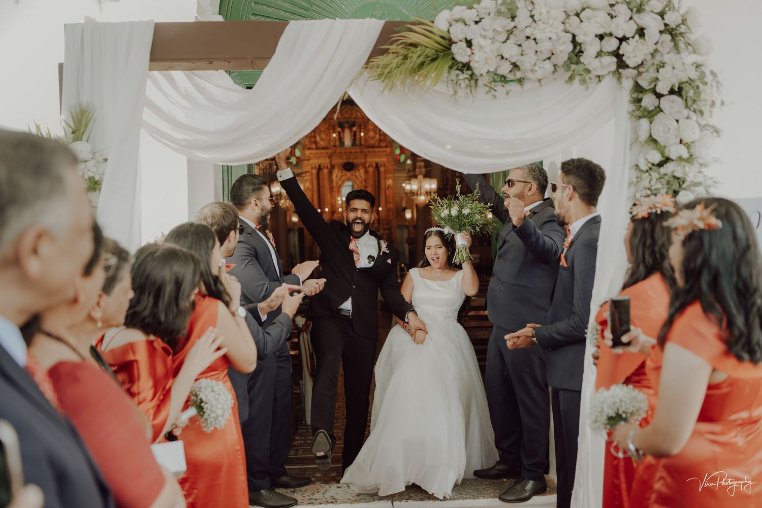 Indoor wedding ceremony with decorative white floral arch and guests in coral colored dresses standing in the aisle.