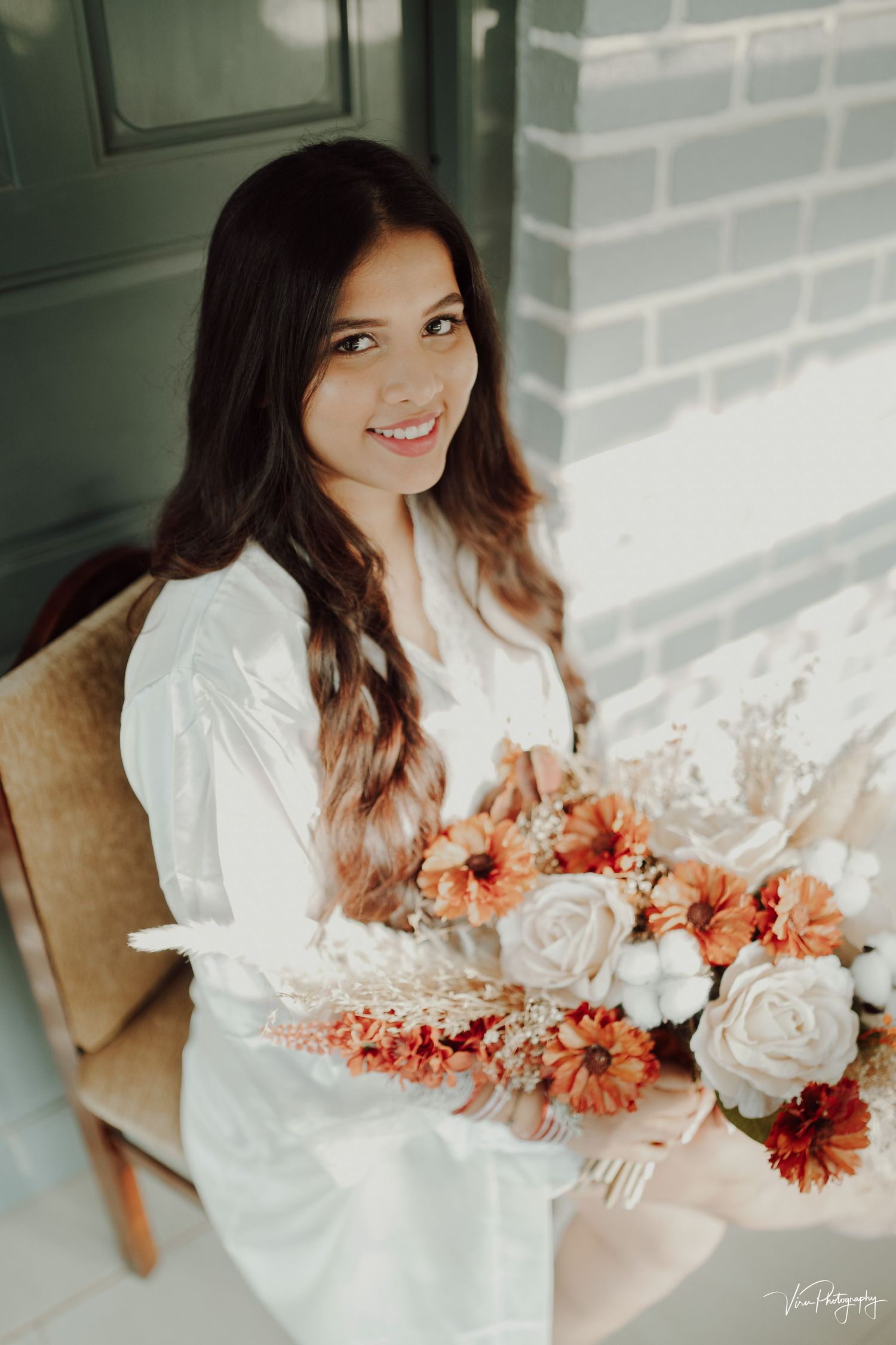 Someone in a white silk robe holds a beautiful bouquet of coral and white flowers while seated on a tan chair.