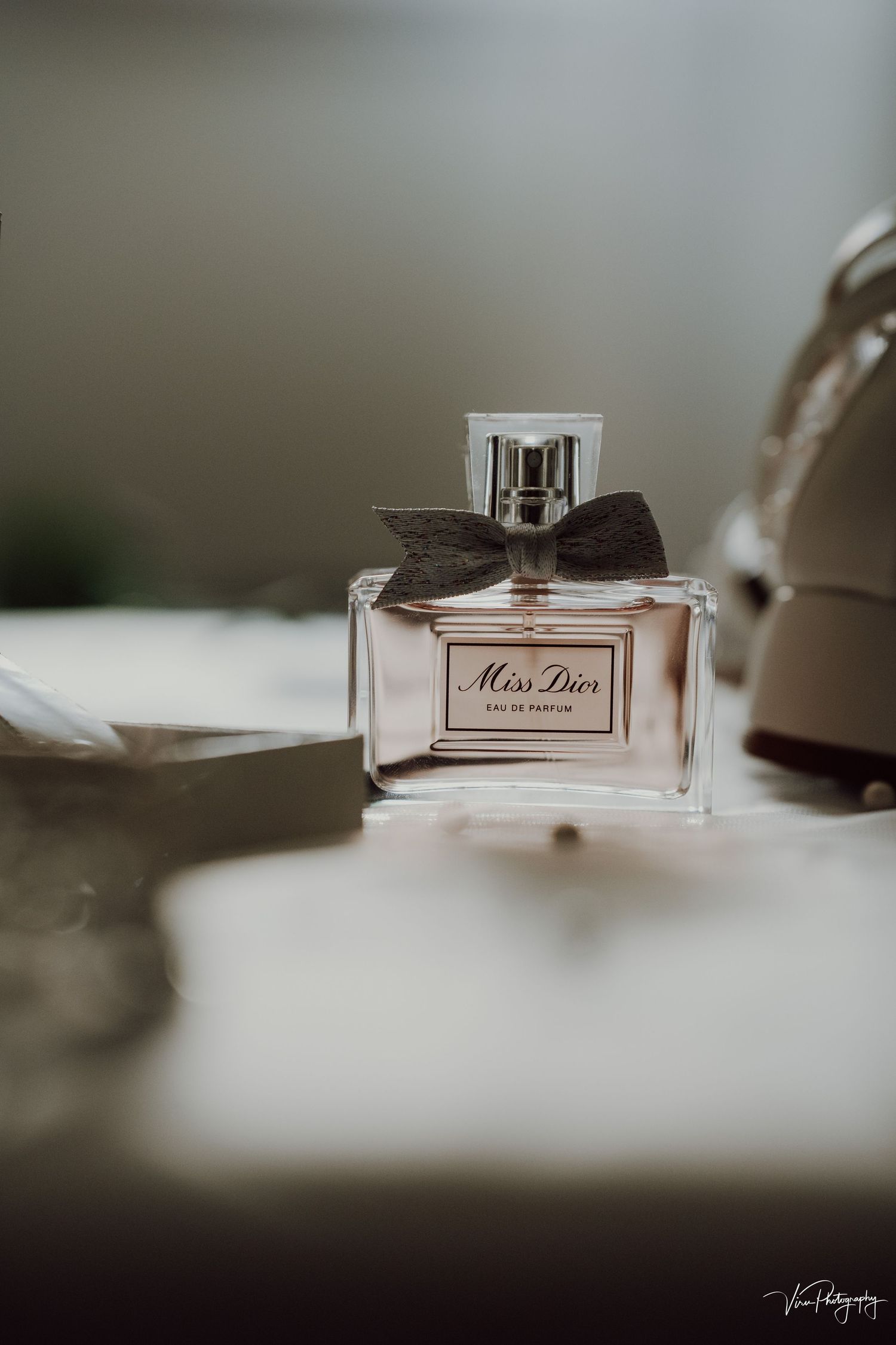 A bottle of Miss Dior perfume with a black bow sits elegantly on a blurred surface in moody lighting.